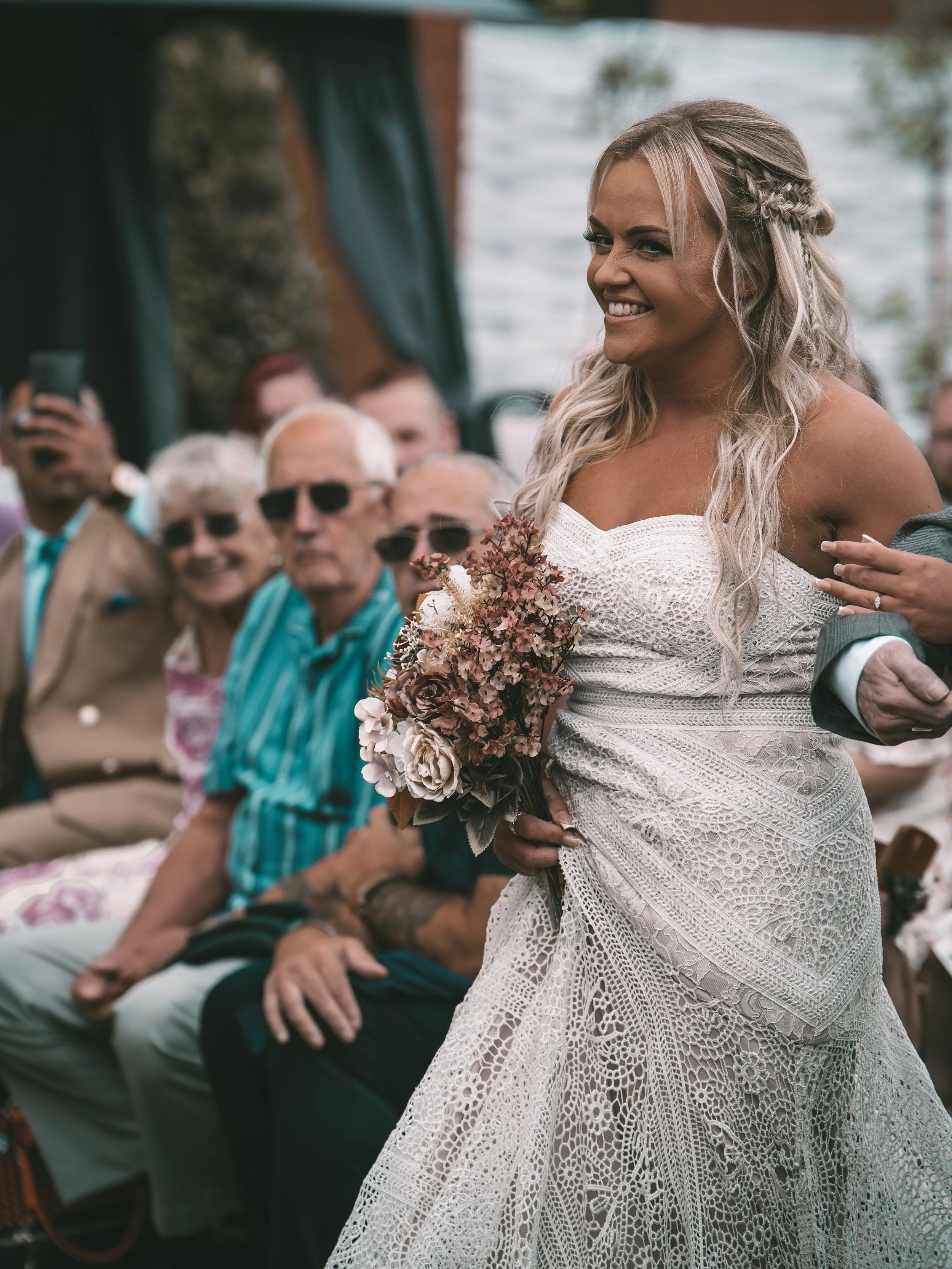 A bride with long, blond hair styled with braids, smiling at her wedding, holding a bouquet of flowers, wearing a white lace wedding dress, with seated guests in the background.