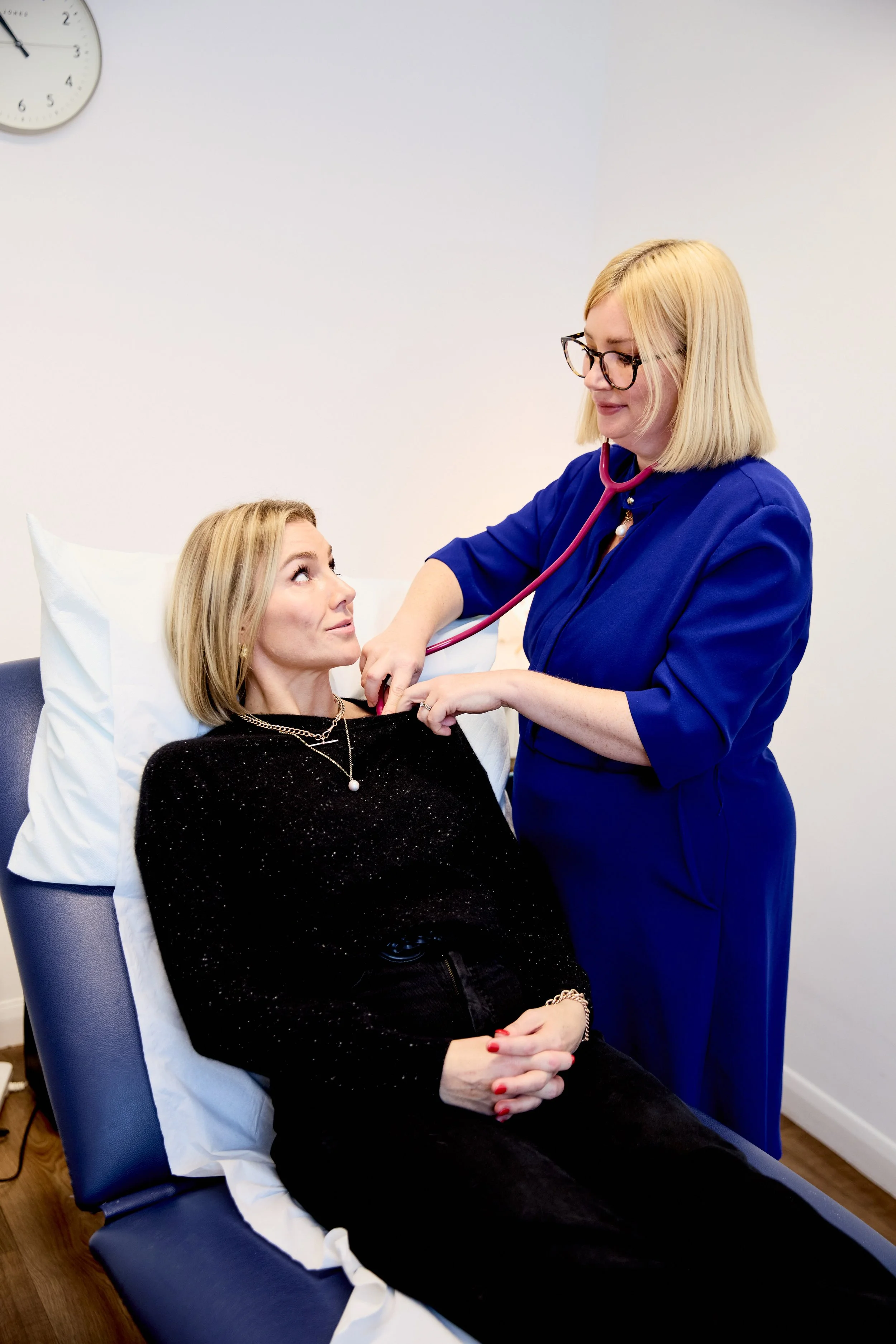Dr Lorraine Kemp wearing glasses and a blue uniform, uses a stethoscope on a woman lying on a medical examination bed. The woman is dressed in black, with blonde hair, and looking up at the healthcare worker.