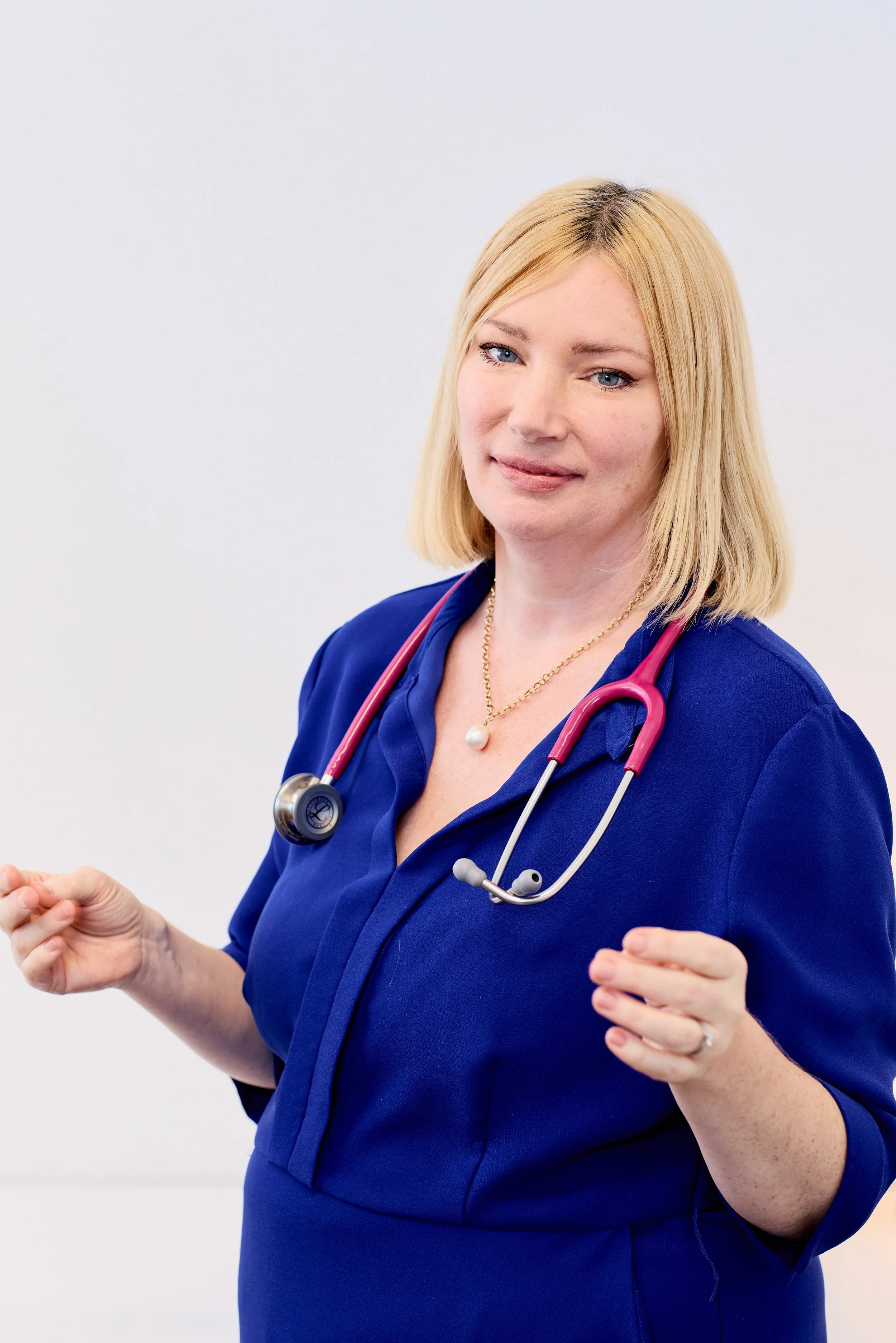 Dr Lorraine Kemp wearing professional wearing a blue medical uniform with a pink stethoscope around her neck, standing against a plain white background.