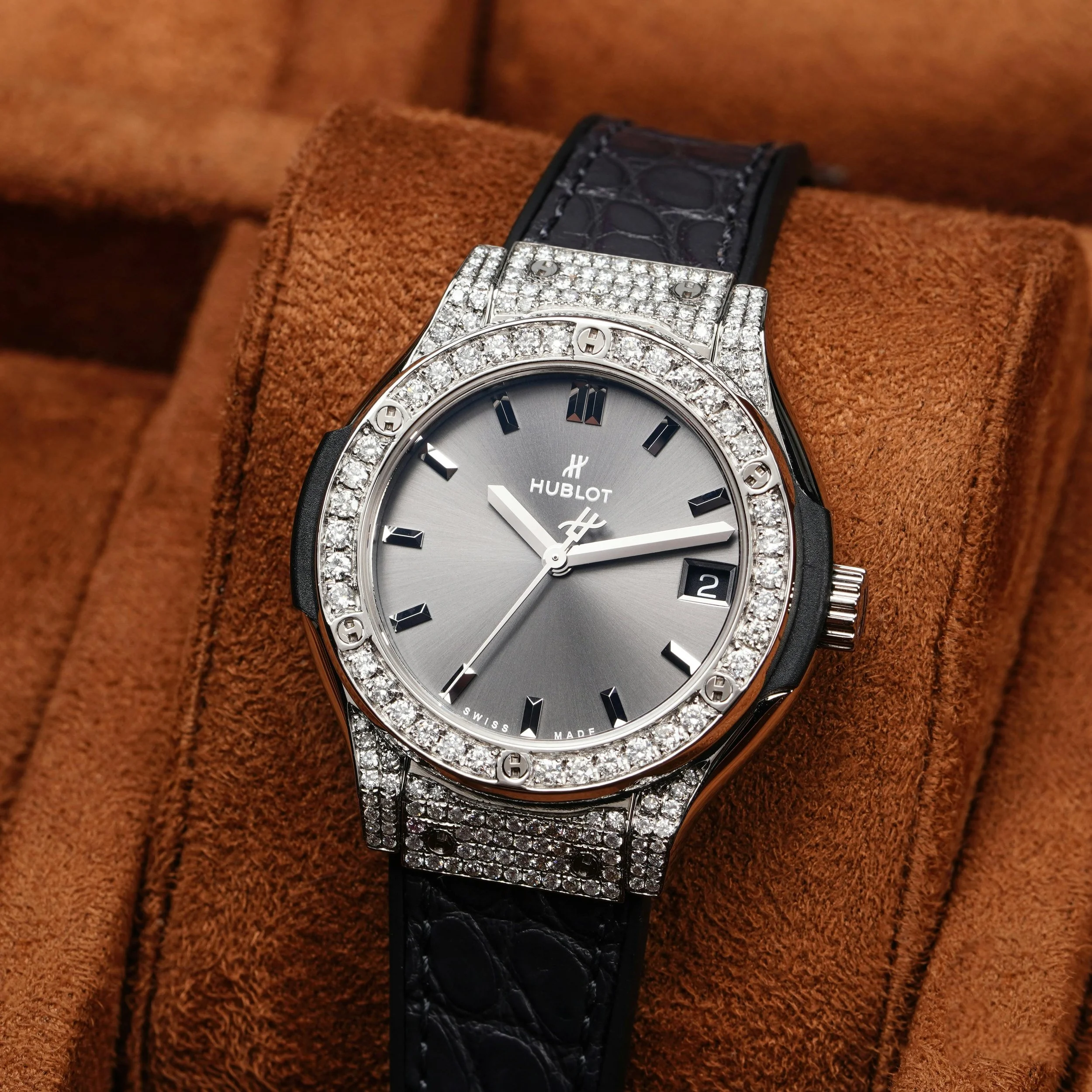 Reloj de lujo Hublot con esfera plateada, bisel y detalles adornados con diamantes, correa de cuero negra, colocada en un soporte de terciopelo marrón.