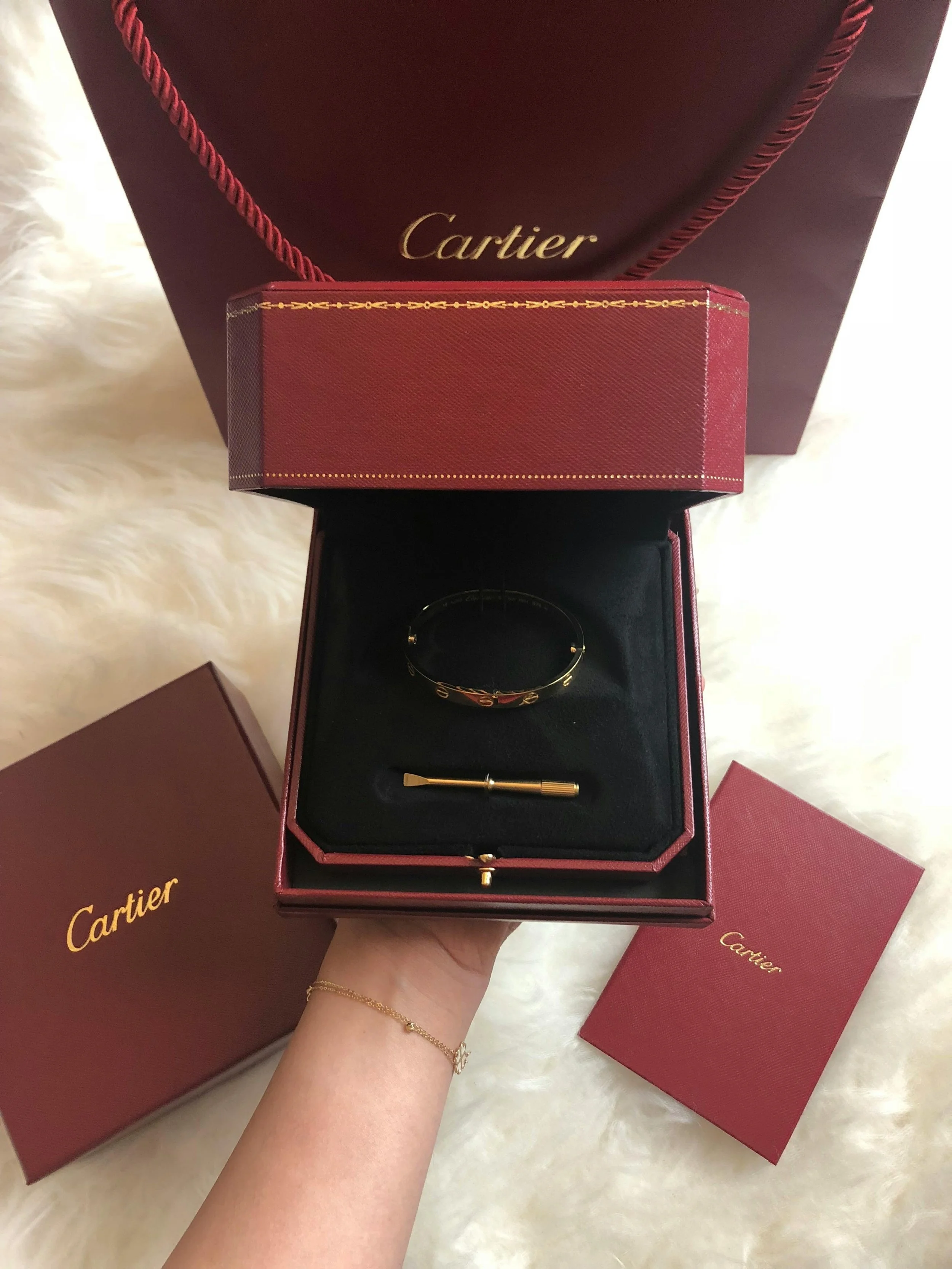 Caja de joyería de Cartier con pulsera y pulsera en el interior, con cajas y bolsa en fondo de color vino.