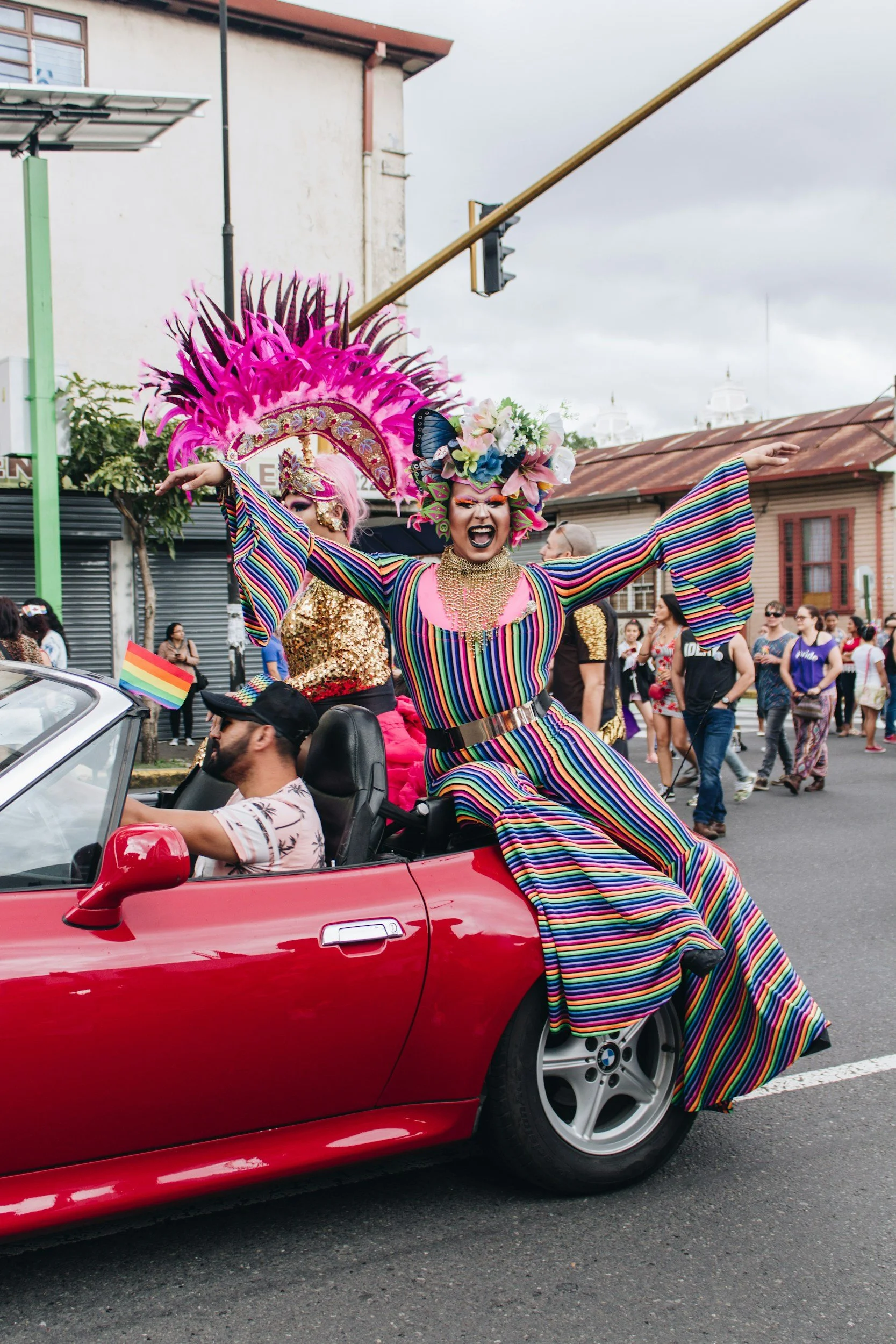 Person in vibrant rainbow-striped dress and elaborate headdress riding in a red convertible during a parade, surrounded by onlookers and supporters.