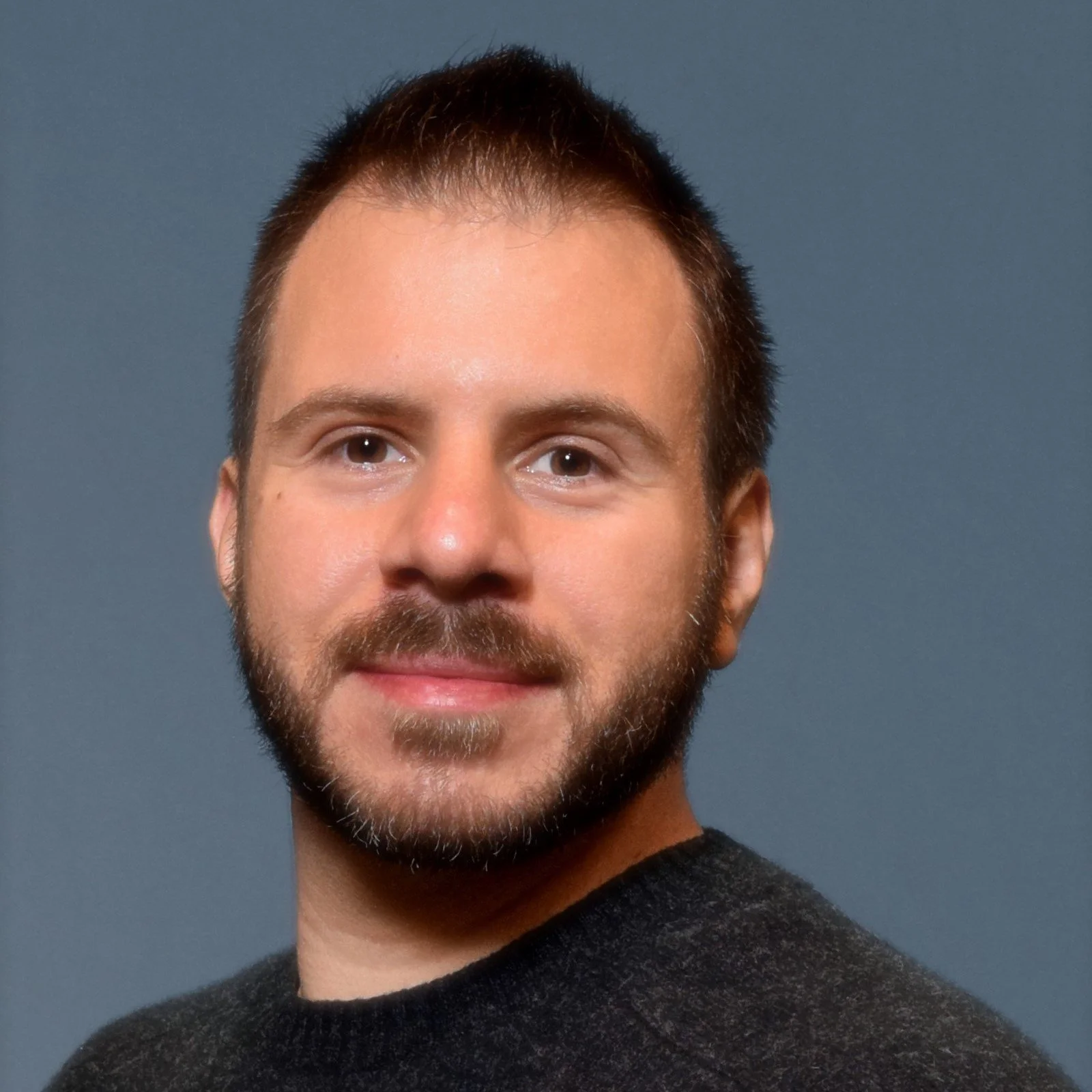 Headshot of a smiling man with short hair, a beard, and wearing a dark sweater against a blue background.