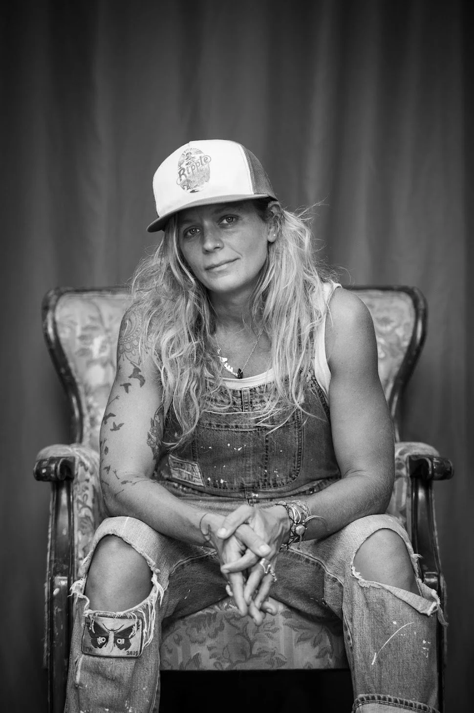 Black-and-white portrait of a person seated in an upholstered chair, leaning slightly forward with hands clasped. They wear a trucker-style cap, a sleeveless shirt under denim overalls, and have long, wavy hair.