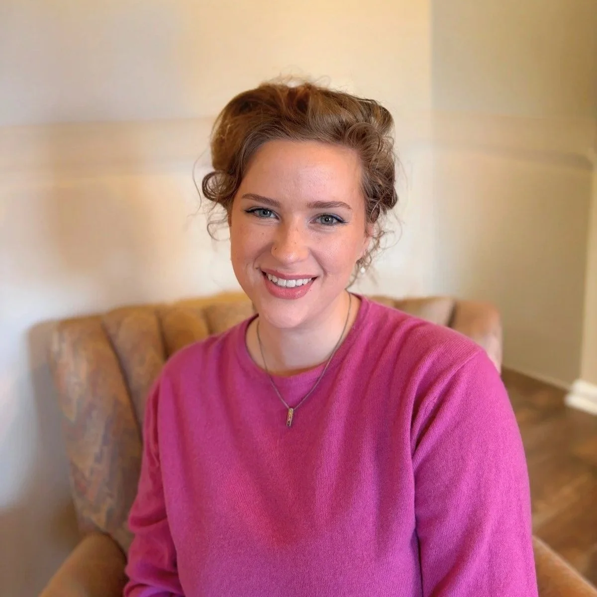 A white femme presenting person with blonde hair, blue eyes, and light makeup wearing a pink sweater, posing against a plain background.