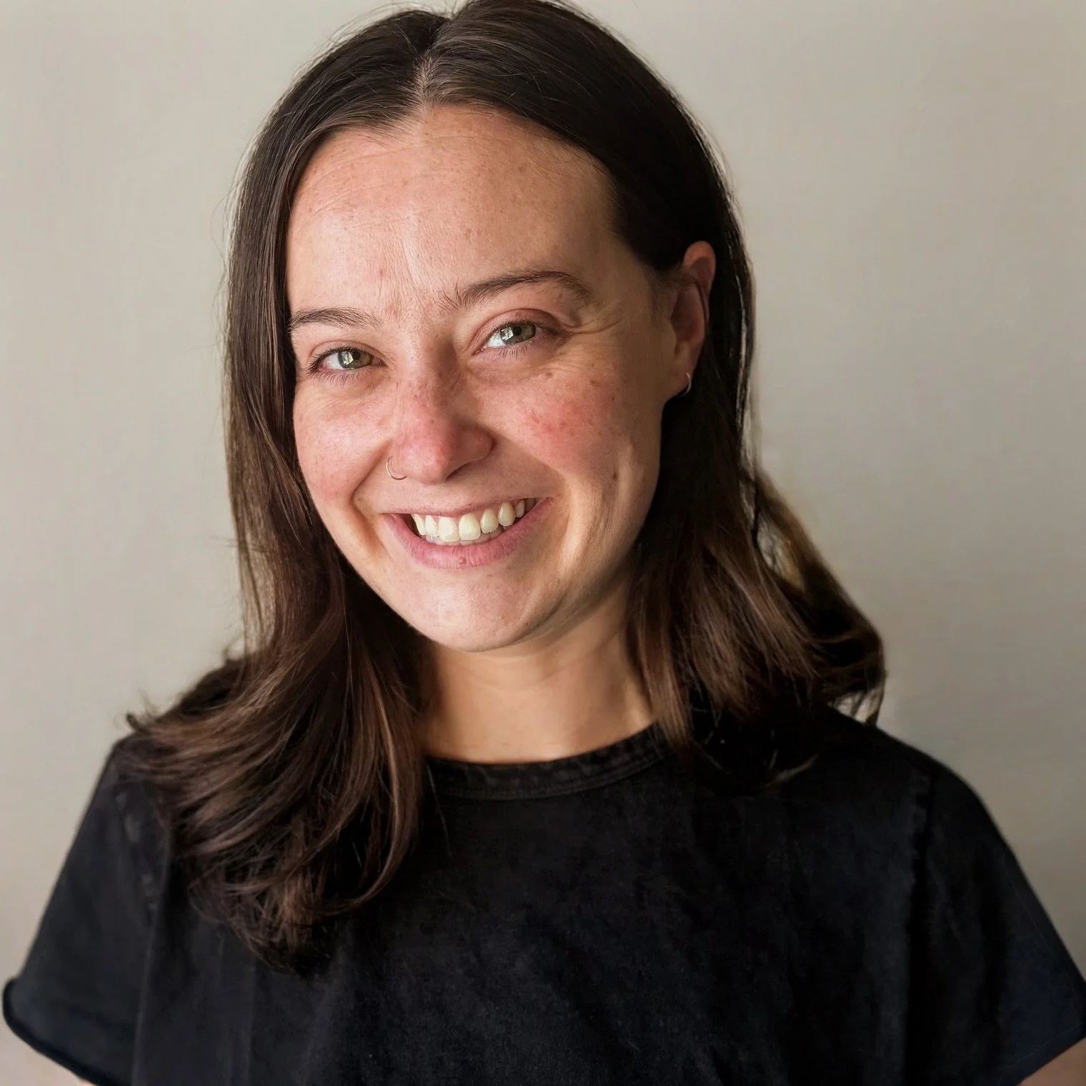 A woman with long brown hair and a nose ring smiling at the camera against a plain light background.