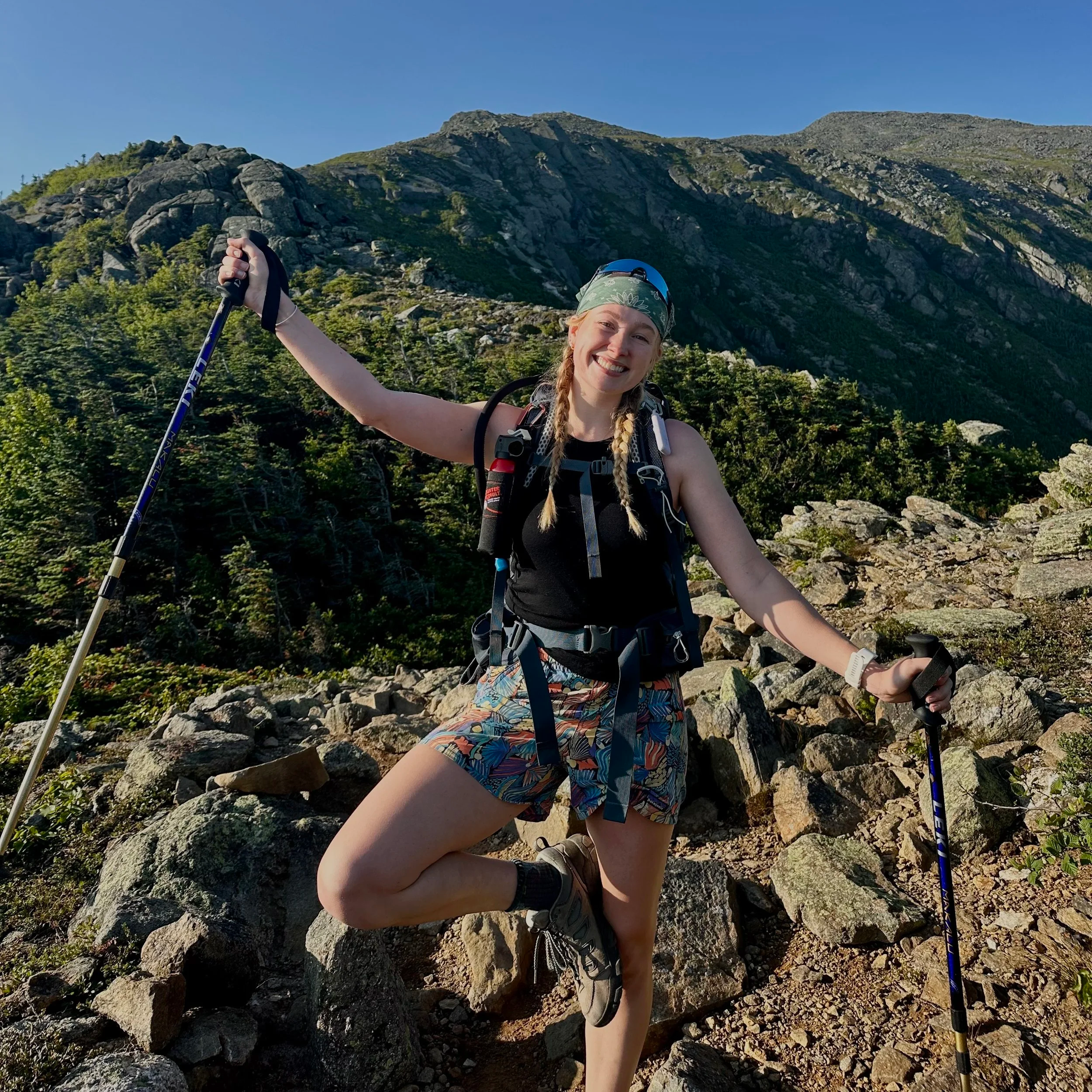 A young woman with blonde hair in braids, wearing a headband, hiking gear, and shorts, standing on rocky terrain with a mountain in the background, smiling, holding hiking poles, and enjoying nature on a sunny day.