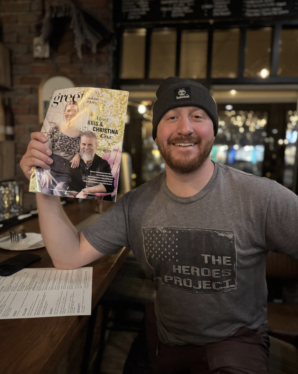 A smiling man with a beard wearing a gray t-shirt and a black beanie, holding a magazine with two people on the cover, in a cozy restaurant or bar setting with warm lighting and a brick wall in the background.