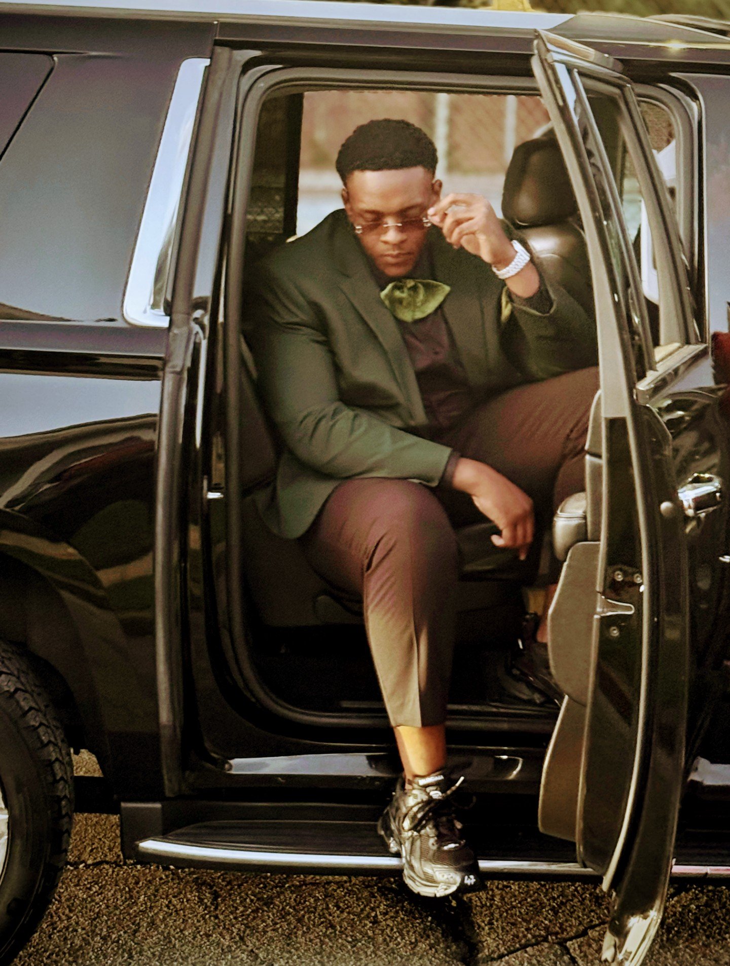 A man sitting inside a black luxury SUV with the door open, adjusting his sunglasses, dressed in a suit with a bow tie, brown pants, and sneakers.
