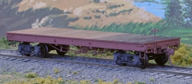Model flatbed train car on tracks with painted landscape background.