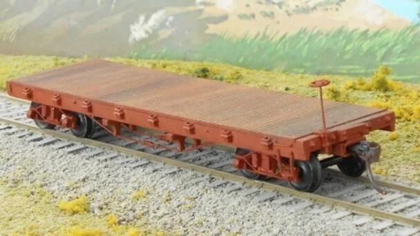 Model flatbed train car on a track with yellow and green landscape background.