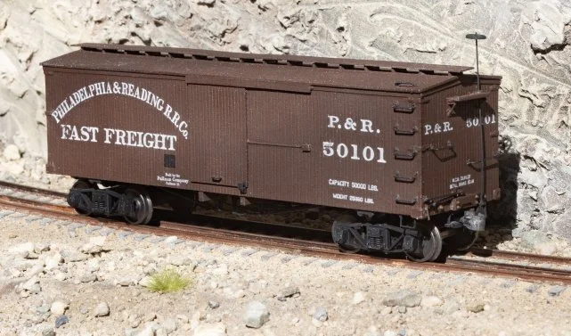 A modern house with wooden and stone exterior walls situated on a green hillside, with snow-capped mountains in the background under a cloudy sky.