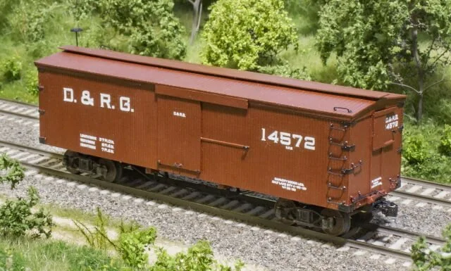 A red boxcar train car marked with D. & R. G., 14572, and other identification numbers, located on railway tracks with green trees in the background.