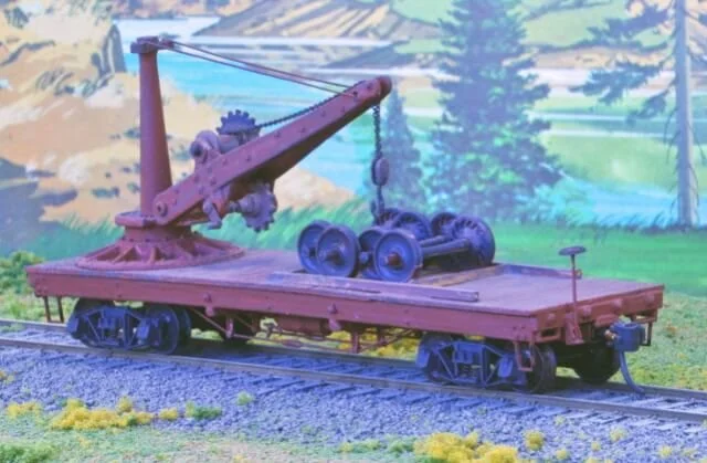 Model of a flatbed train car with a crane and wheels, set on train tracks with a scenic backdrop.