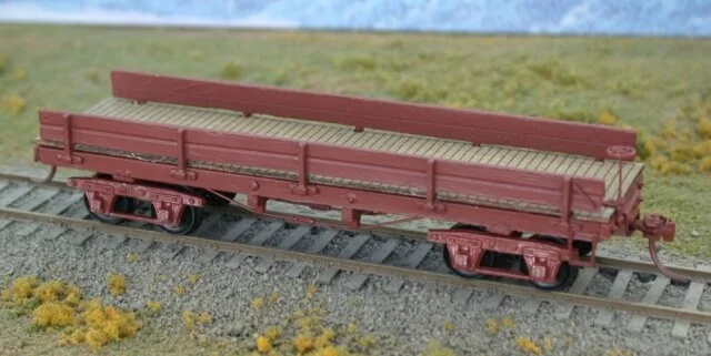 A model train flatbed car on a railway track with grass and landscape in the background.