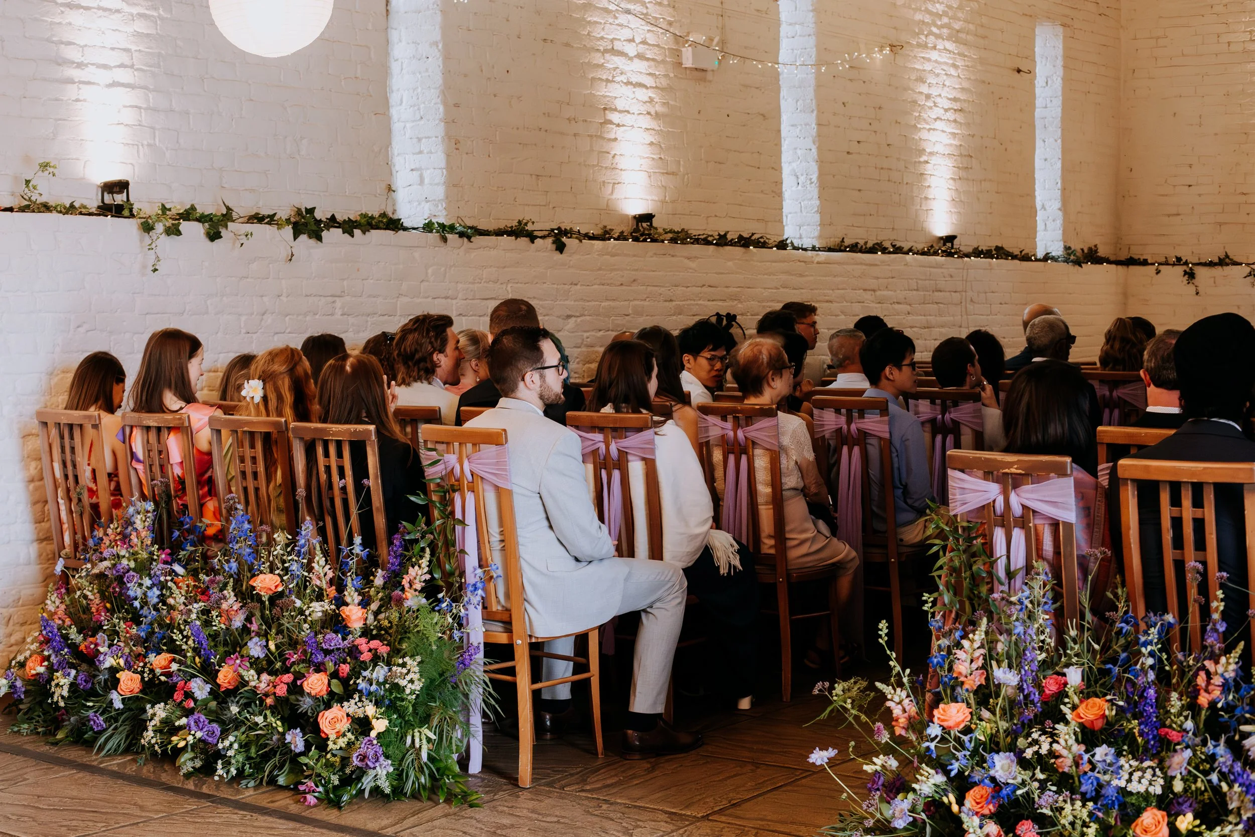 A wedding ceremony is taking place with colourful meadows of flowers decorating the back row of chairs. 