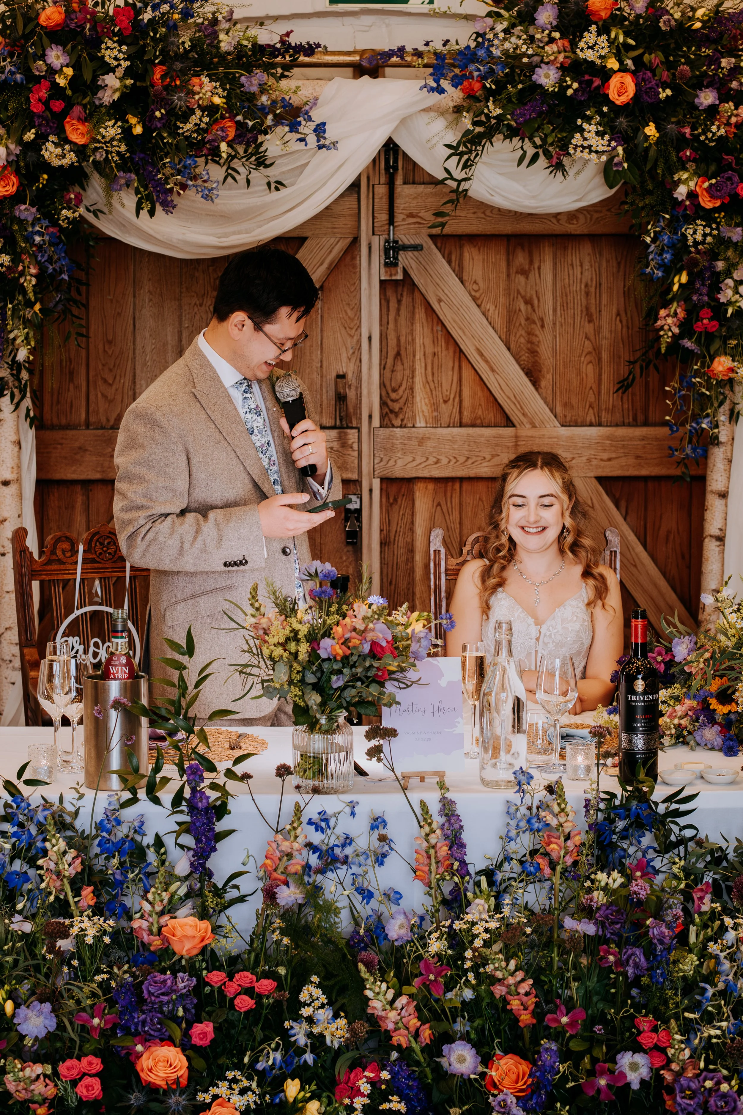 A groom making his speech at the top table surrounded by his bride and many colourful flowers. 