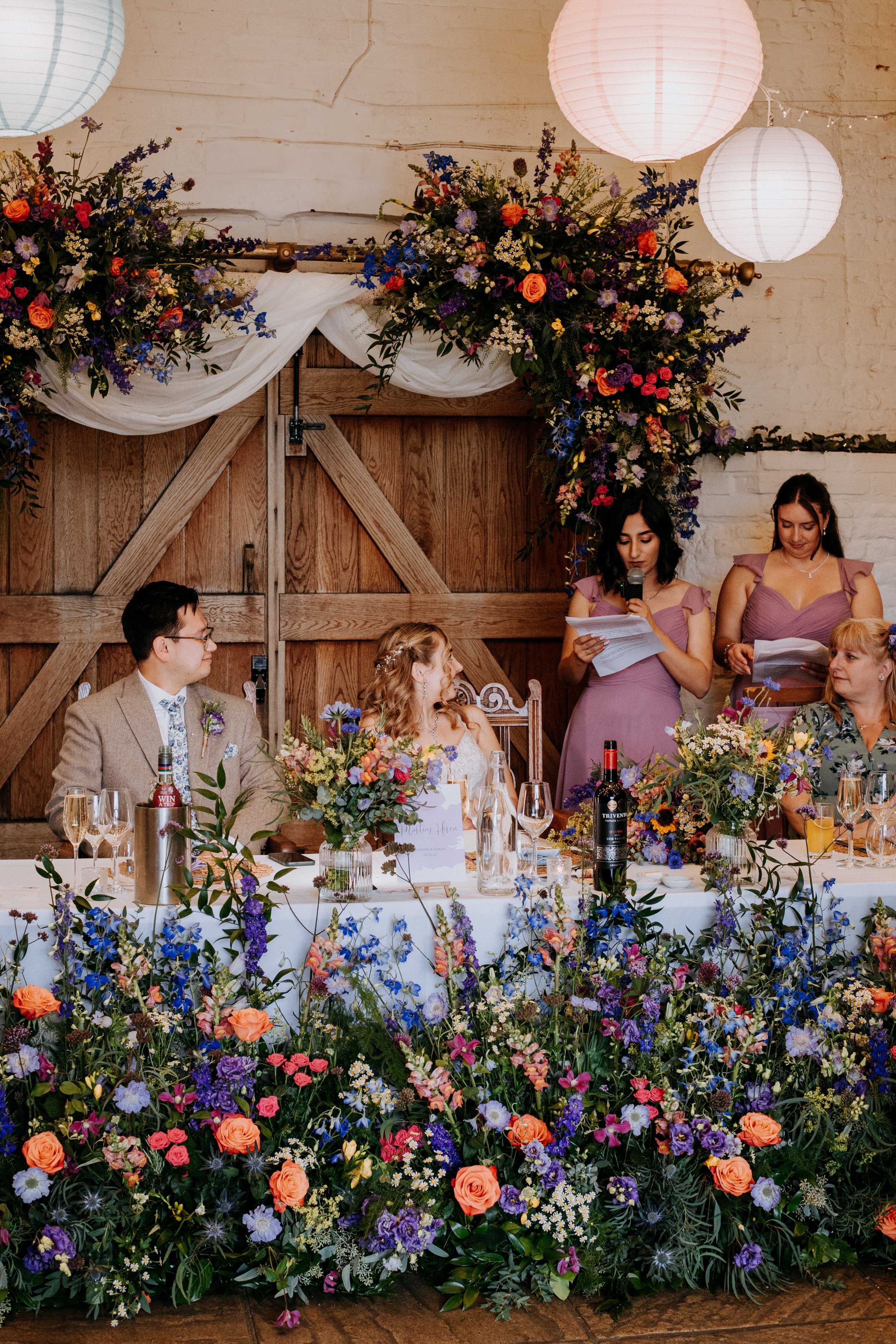 A bride and groom are sat at their top table surrounded by guests making speeches. There are meadows of flowers decorating the front of the top table and a colourful flower arch is behind them. 