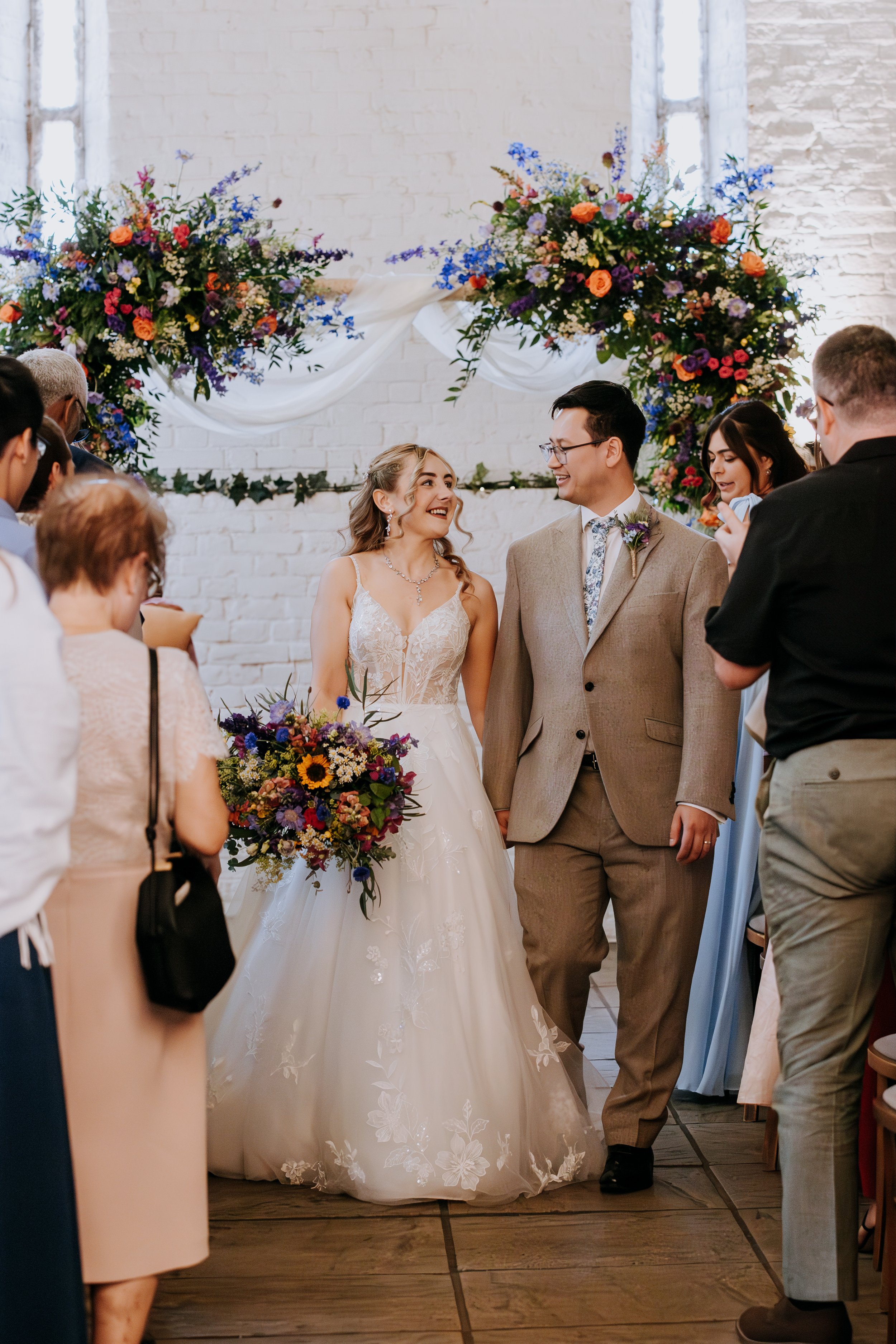 A bride and groom making their way down the aisle after their ceremony. A colourful arch of flowers surrounds them 