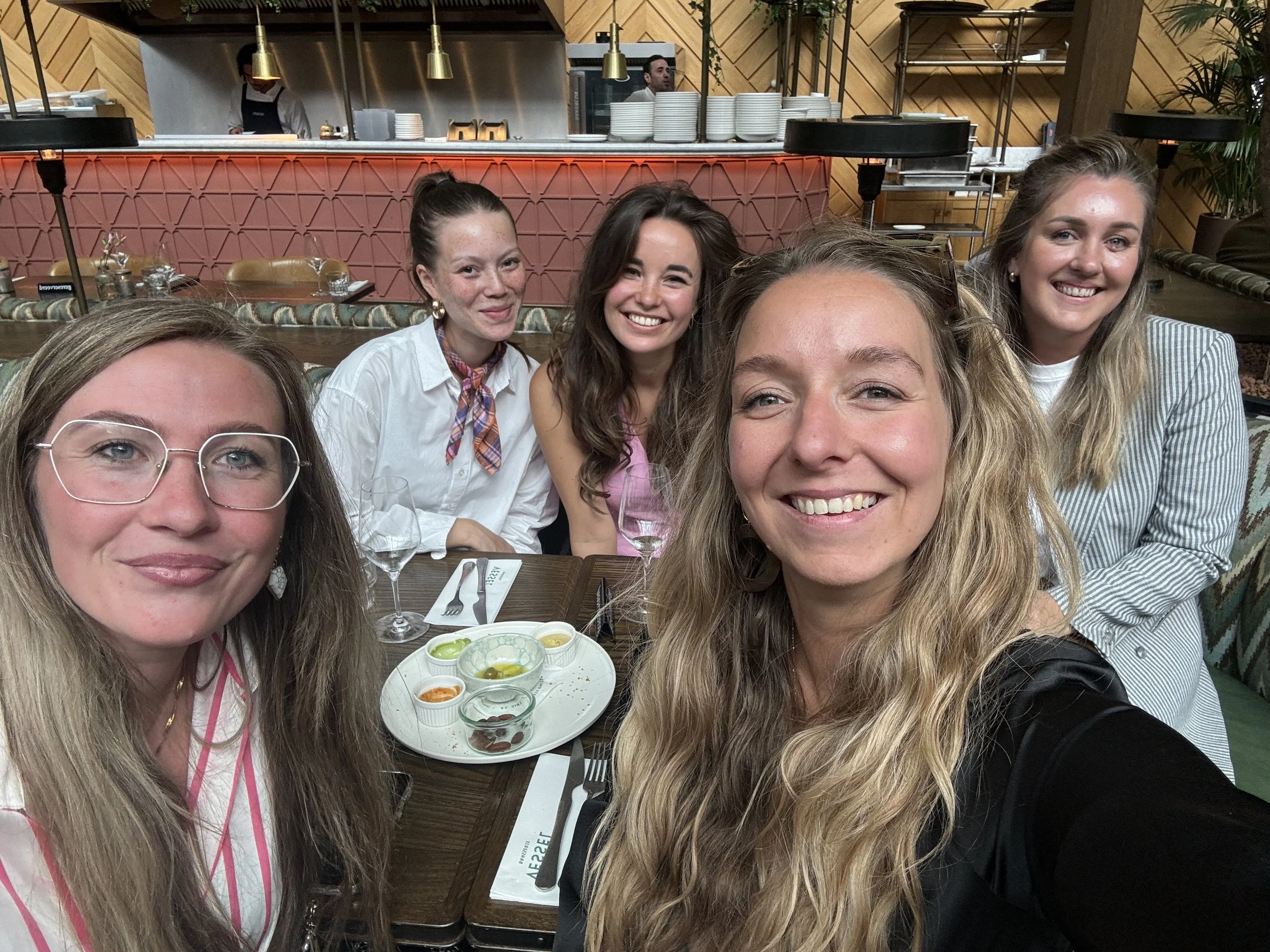 Vijf vrouwen die samen genieten in een restaurant, met een serveerster op de achtergrond en een tafel met ruccola en saus.