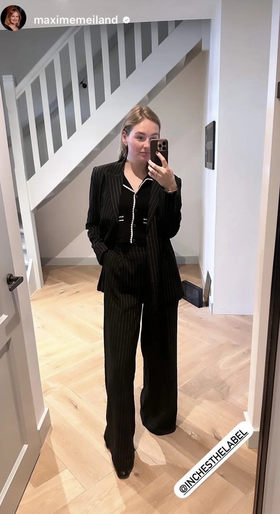 Vrouw in een zwarte broek en blazer met witte strepen, staand voor een spiegel, neem selfie met haar telefoon, in een interieur met houten vloer en witte muur, trappen op de achtergrond.