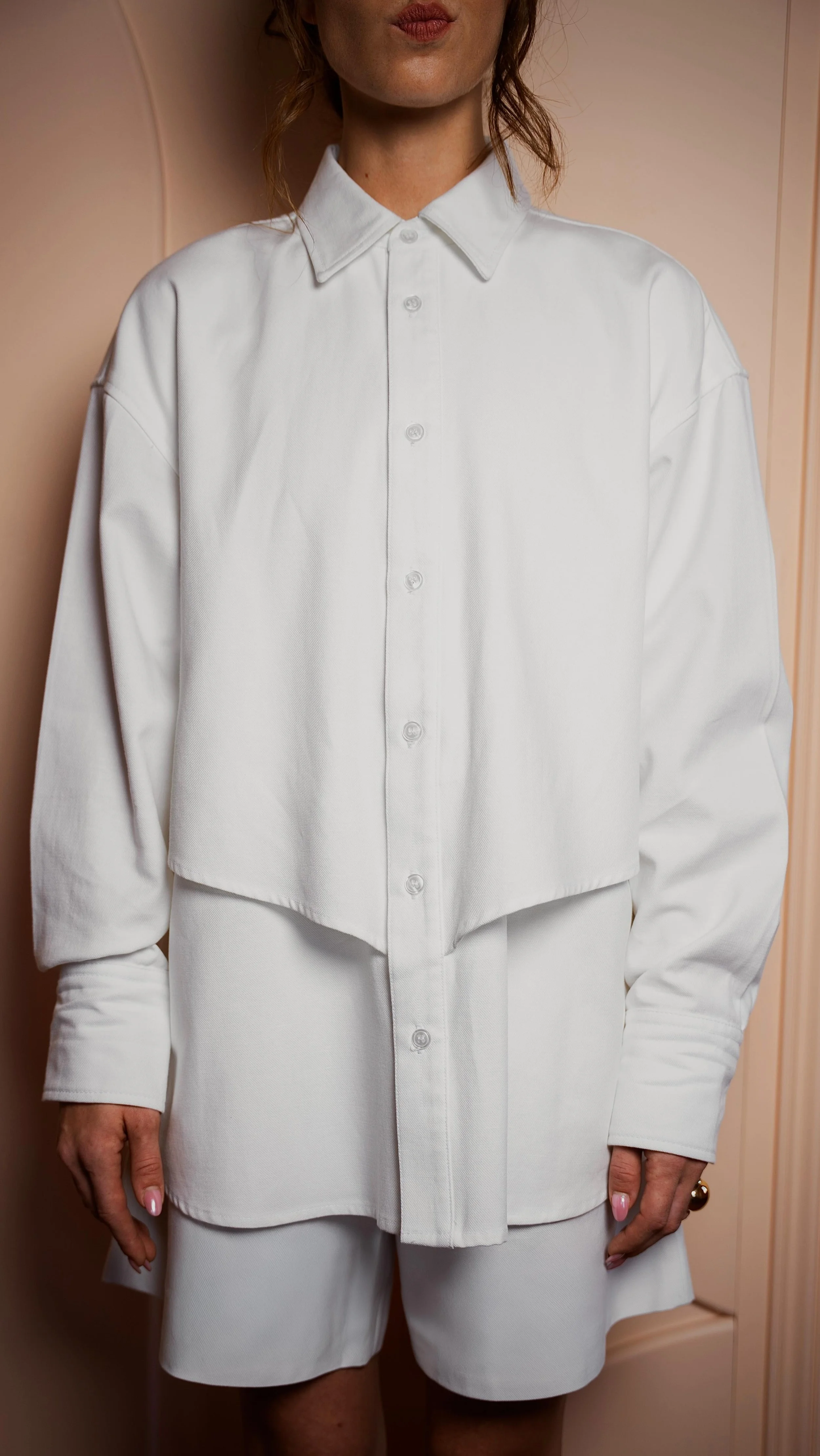 Close-up van een vrouw die een witte overhemd en bijpassende witte short draagt, standing voor een lichtroze muur.