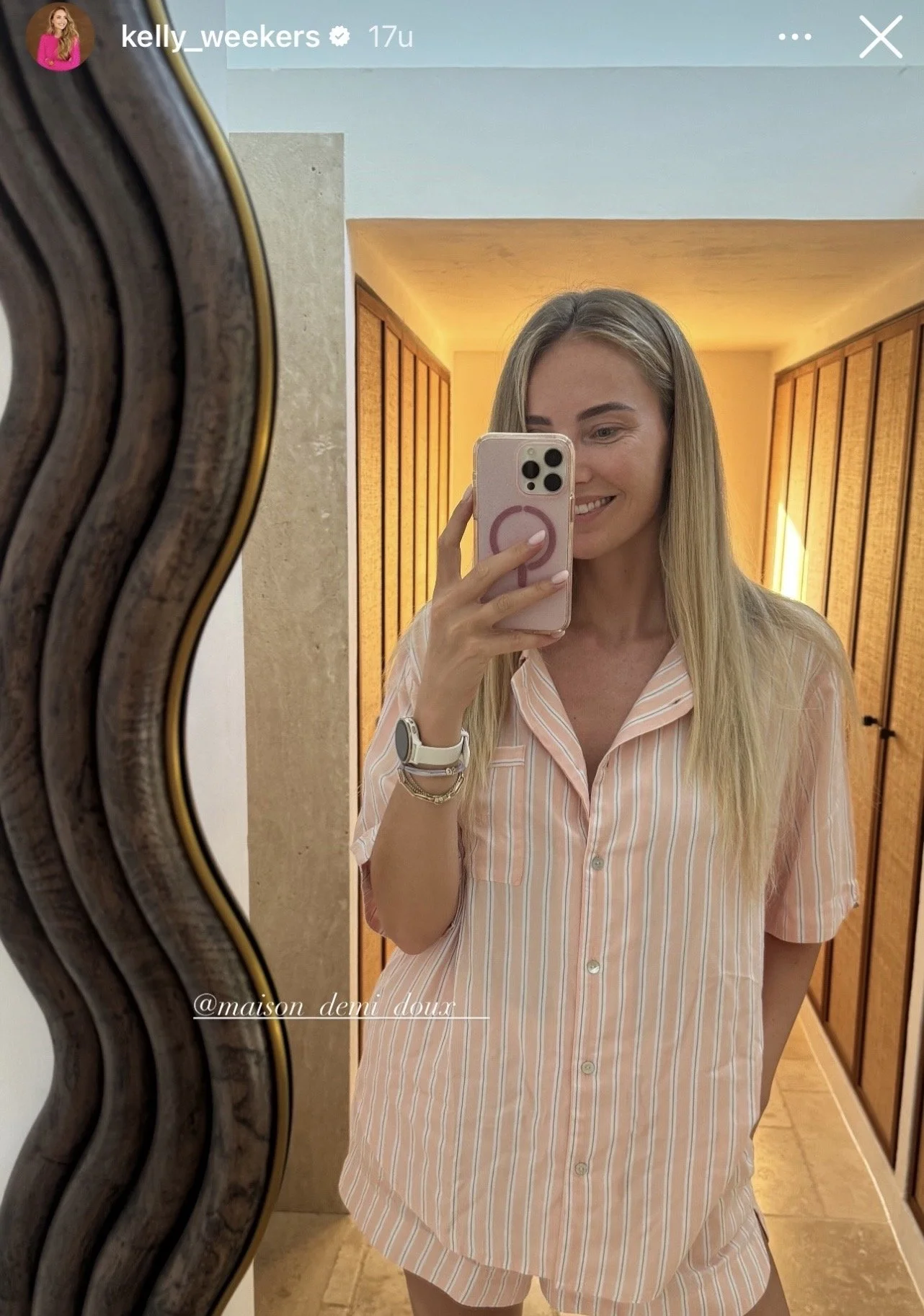 Vrouw maakt een selfie in een spiegel, ze draagt een roze en witte gestreepte pyjama, met een witte smartwatch aan haar pols. Achter haar bevinden zich houten kleedkamers en een warme verlichting.