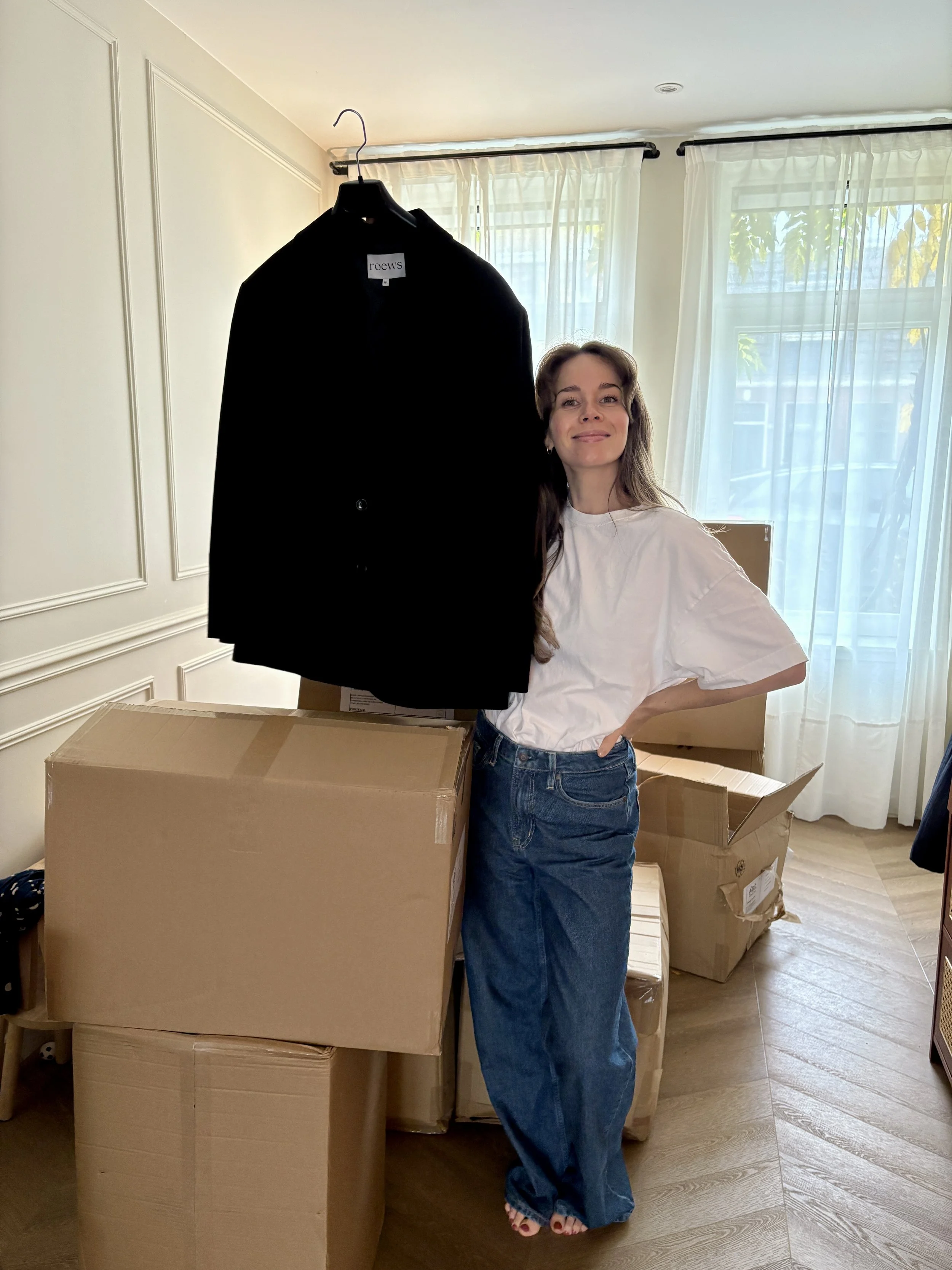 Vrouw in witte T-shirt en grote blauw jeans poseert naast een grote kartonnen doos in een kamer met grote ramen en witte gordijnen, omgeven door meerdere gesloten kartonnen dozen.