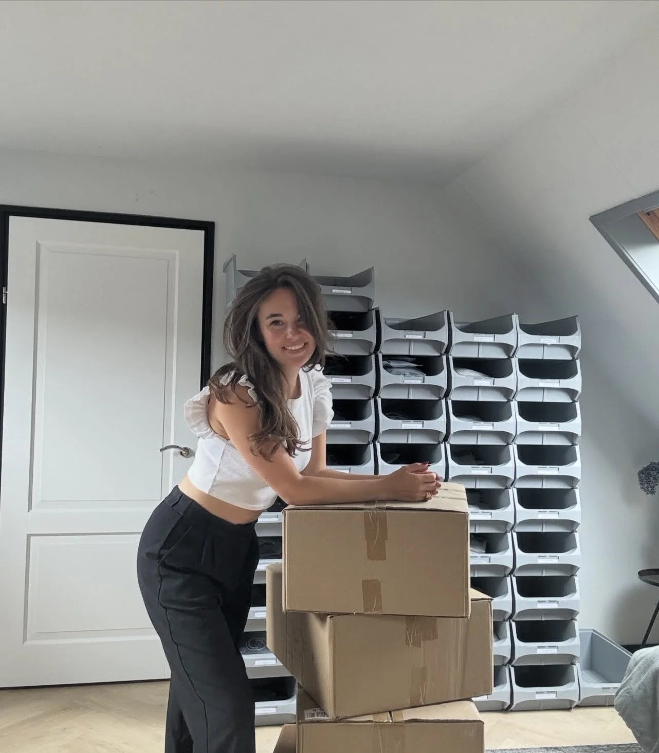 een lachende vrouw met lang, bruin haar die achter een stapel kartonnen dozen staat in een kamer met opbergvakken op de achtergrond.