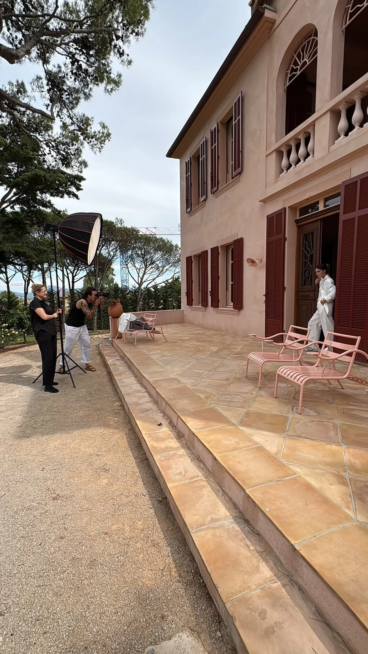 Fotoshoot op het terras van een huis. Twee fotografen maken foto's van een vrouw in een witte pak die op de deurpost van het huis staat. Er is professionele belichting en tuinmeubilair op de patio.