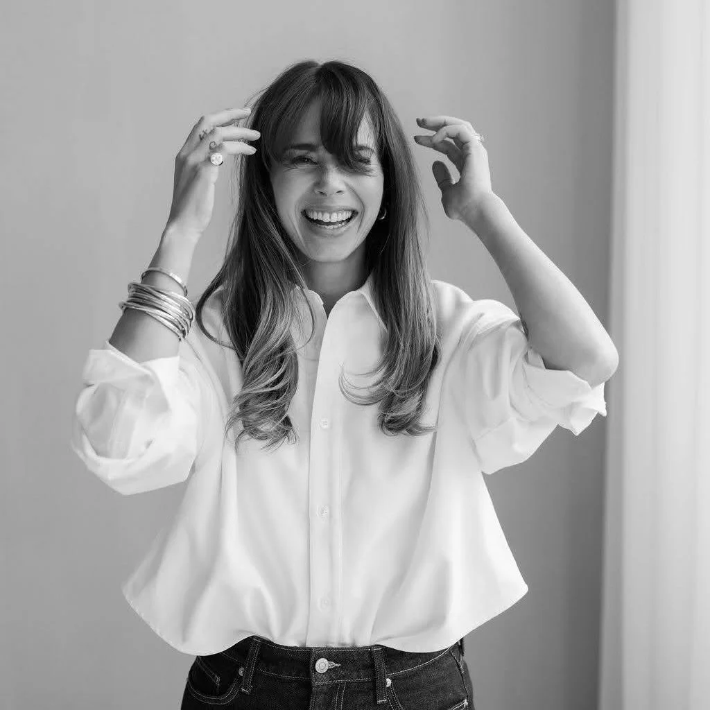 Een lachende vrouw met lang haar en meerdere armbanden, die haar haar kamt of stylet, in een lichte blouse en jeans, binnen, zwart-wit foto.