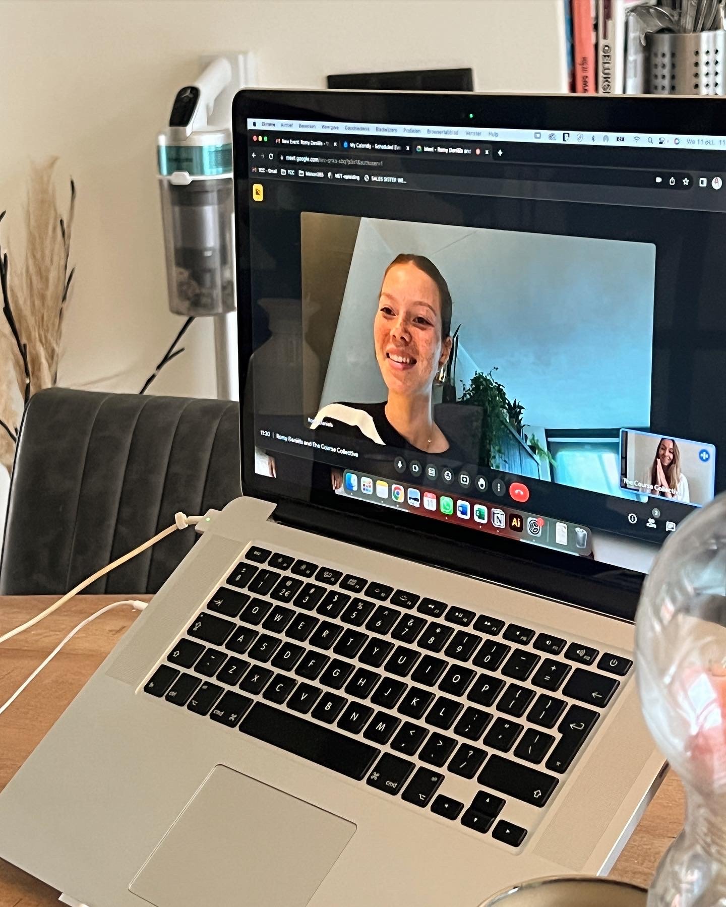 Een laptop op een tafel met een videooproep waarin twee vrouwen zichtbaar zijn, één met een zwarte en witte trui en de ander met een witte trui. Op de achtergrond staat een plant en een raam.
