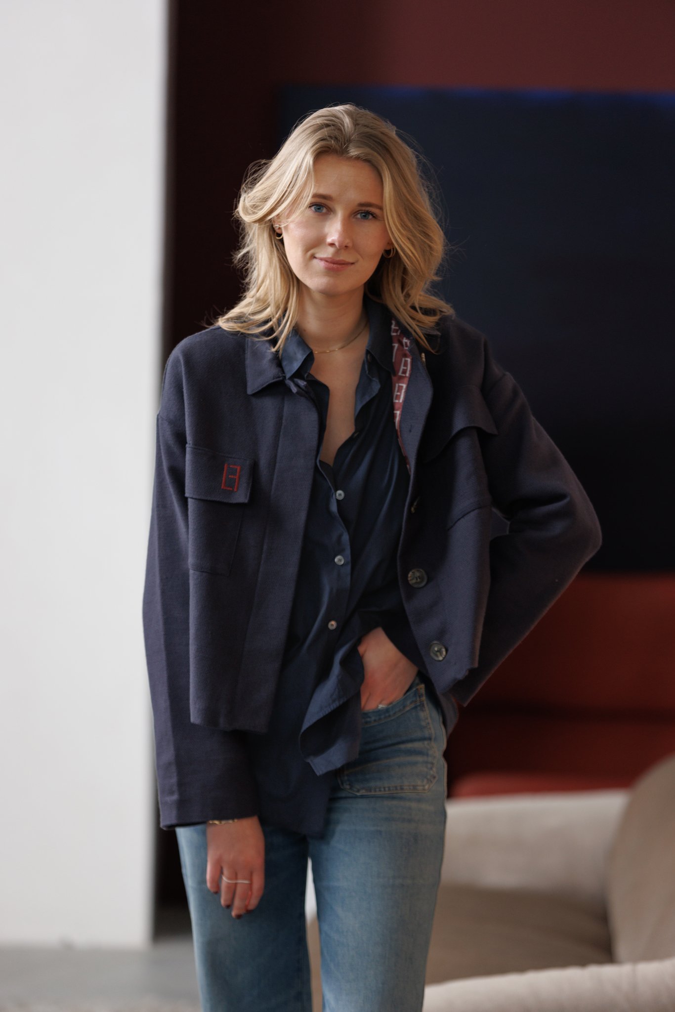 Een vrouw met blonde haar draagt een blauwe blouse en jas, en heeft een hand in haar broekzak. Ze staat binnen, mogelijk in een woning of kantoor.