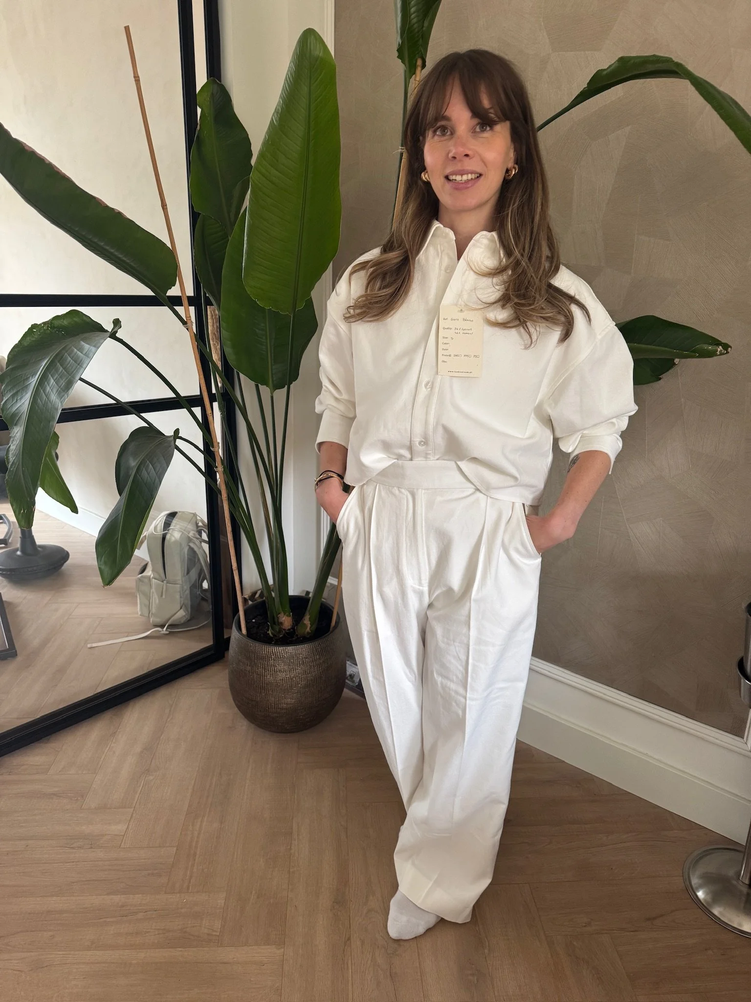 Vrouw met lang bruin haar, witte blouse en beige broek, staand naast een grote groene plant in een kamertje met houten vloer.