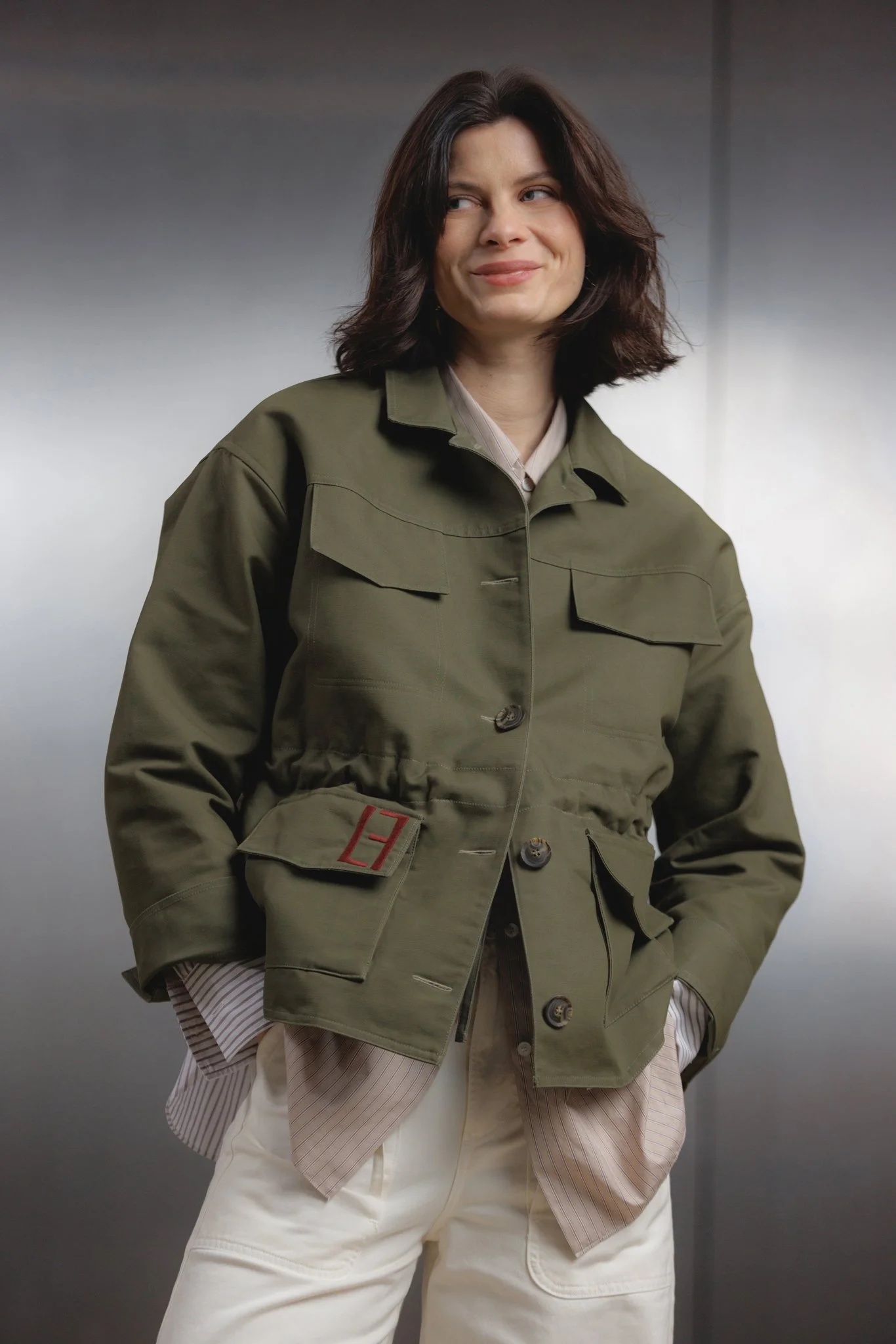 Een vrouw met schouderlang donker haar draagt een groene jas en losse beige broek, poserend voor een effen achtergrond.