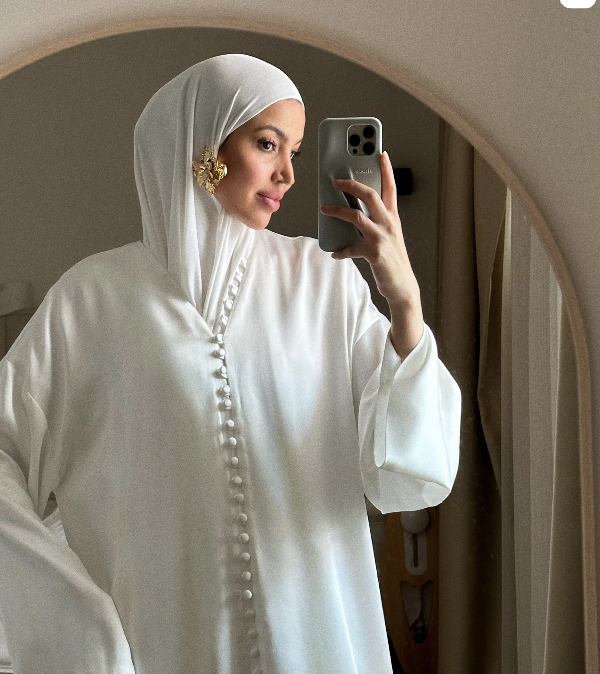 Een vrouw draagt een witte hijab en een witte lange jurk, terwijl ze een spiegelselfie neemt met haar smartphone.