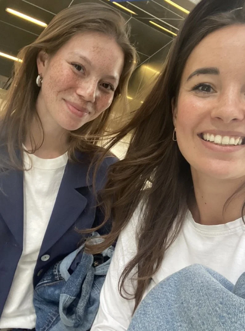Twee jonge vrouwen die vrolijk glimlachend een selfie maken, in een interieur met donkere muren en gele lichteffecten.