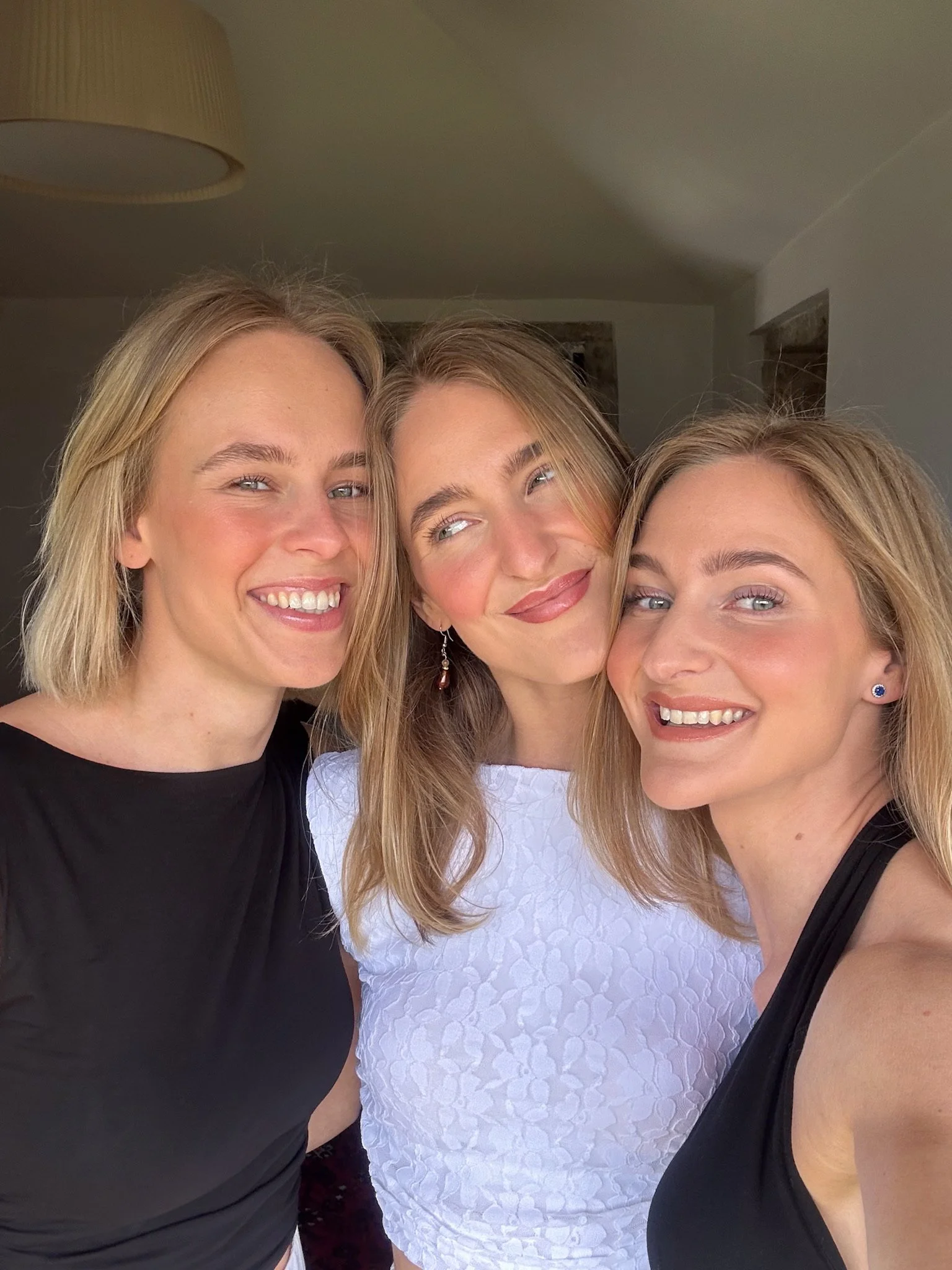 Drie vrouwen die dicht bij elkaar staan en glimlachen, met een relaxte achtergrond van een interieur.
