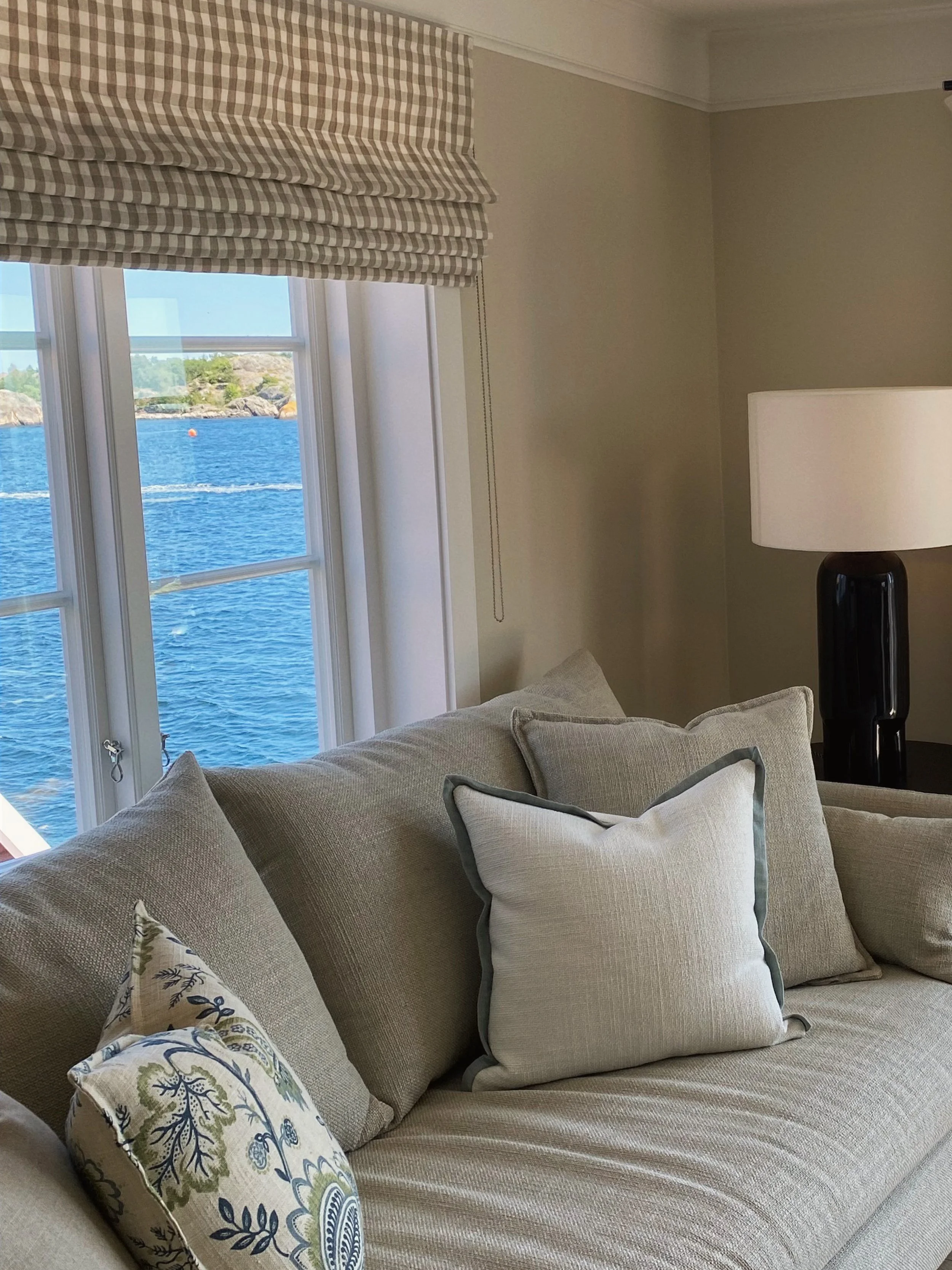Living room with beige sofa, various pillows, a window with a curtain, and a lamp, overlooking water and coastline outside.