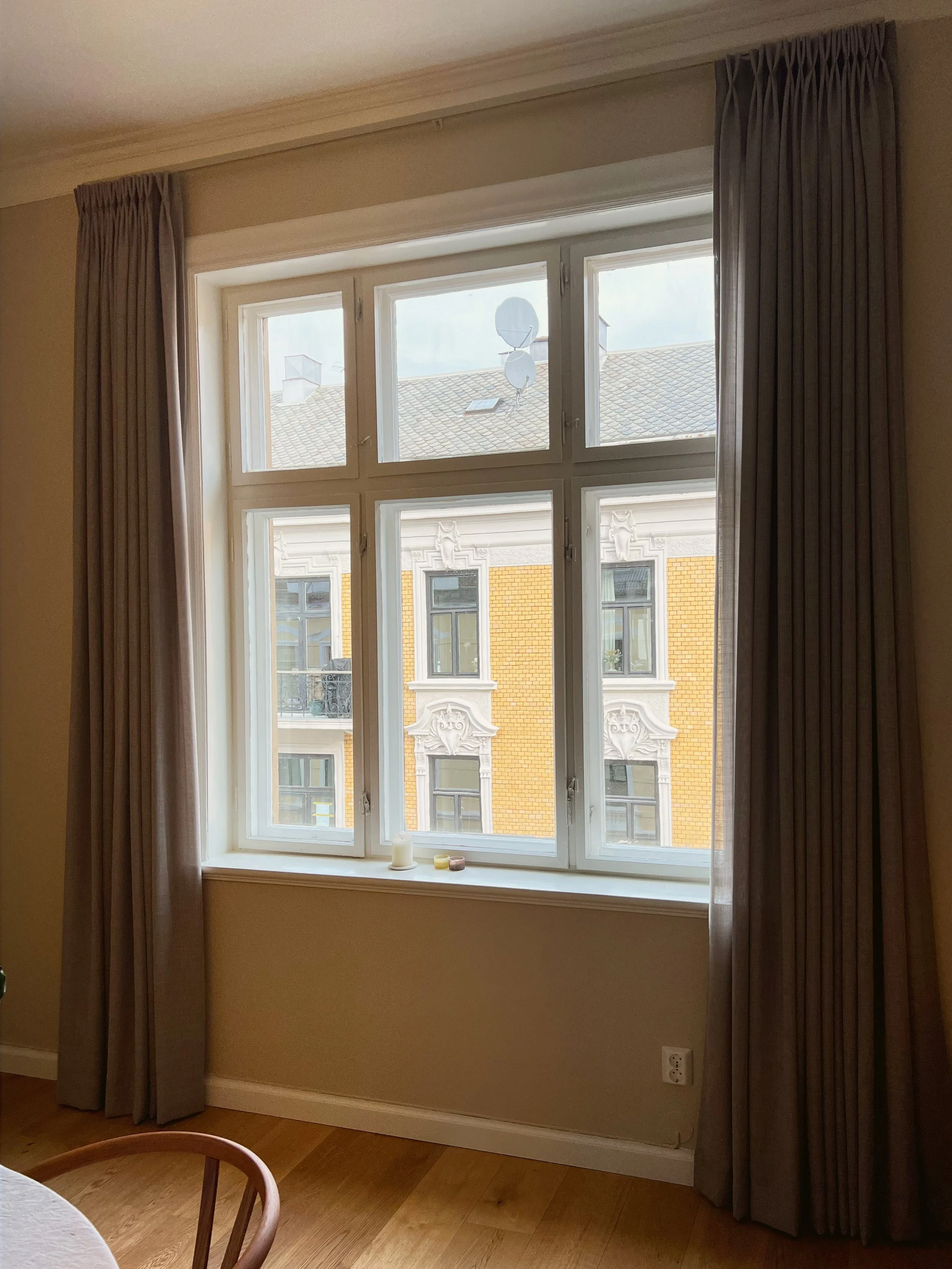 A large window with white frame and multiple panes, flanked by beige curtains, overlooking a building with ornate architectural details and a yellow brick facade, with candles on the windowsill.