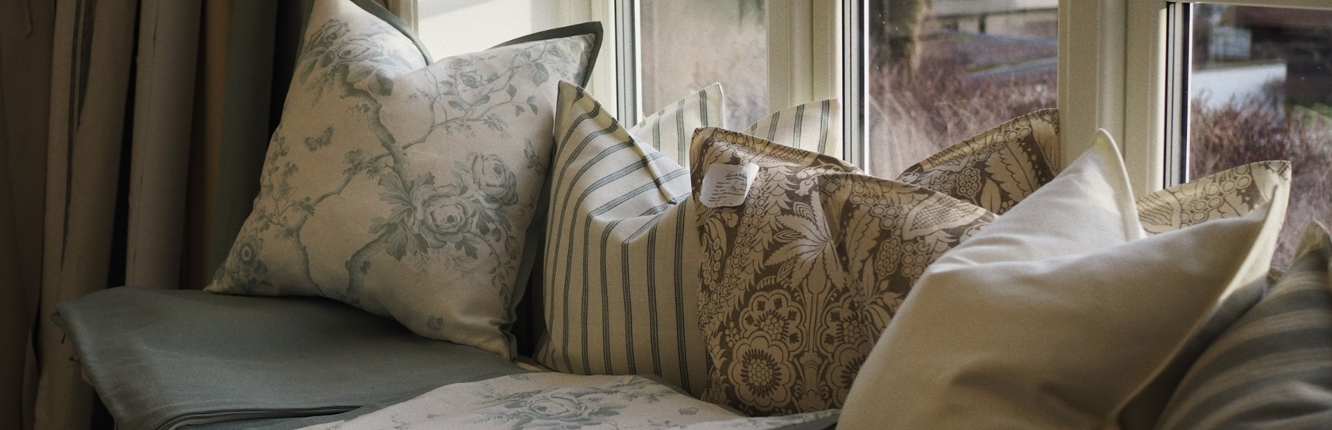 A window seat with several decorative pillows with floral and striped patterns, some with tags, arranged on a window sill, overlooking an outdoor scene with trees and a sidewalk.