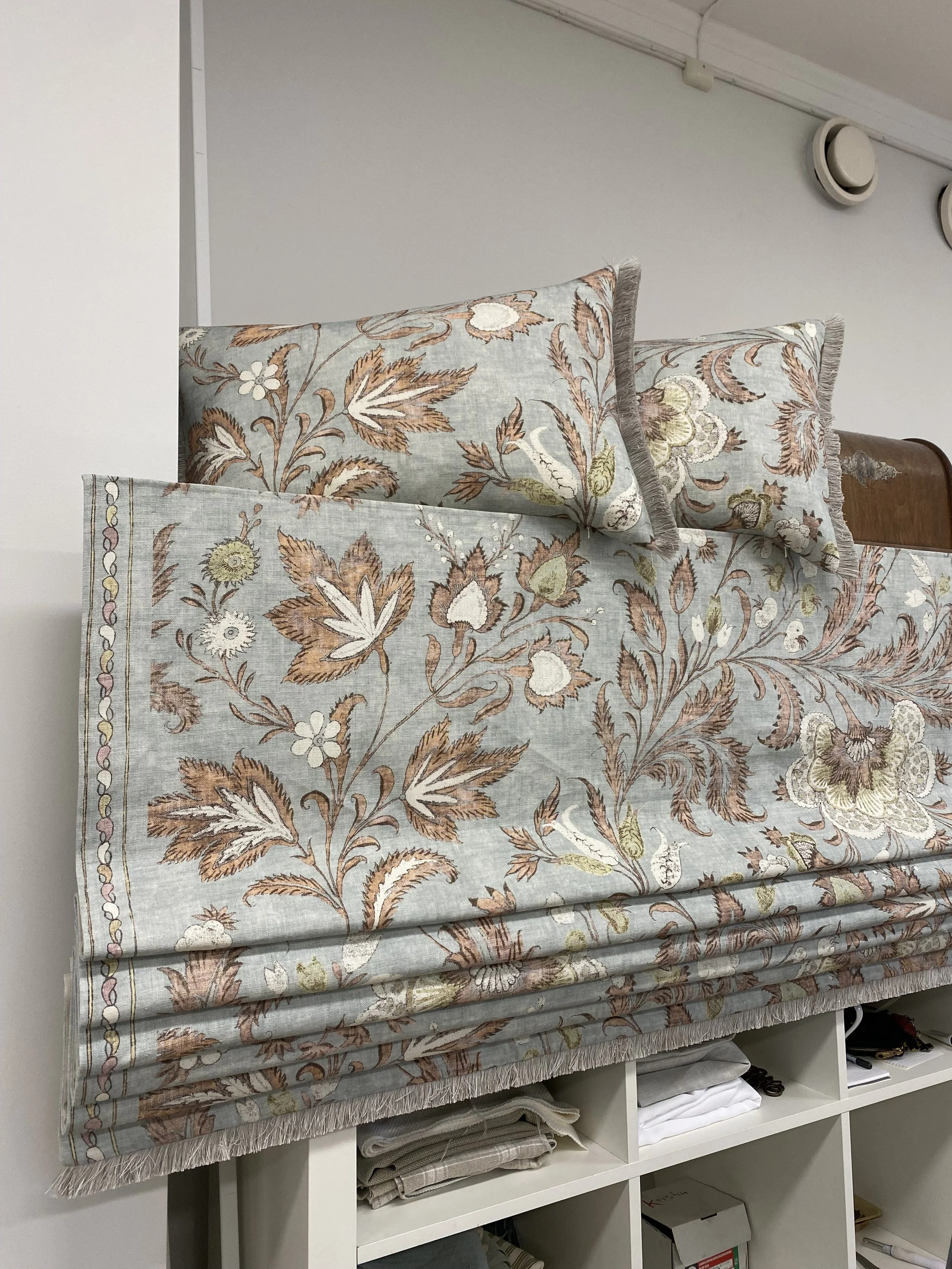 A set of embroidered curtains with floral pattern are hanging in front of a white shelf filled with folded towels, boxes, and miscellaneous items. The curtains have a pastel blue background with brown, beige, green, and white floral embroidery, and f