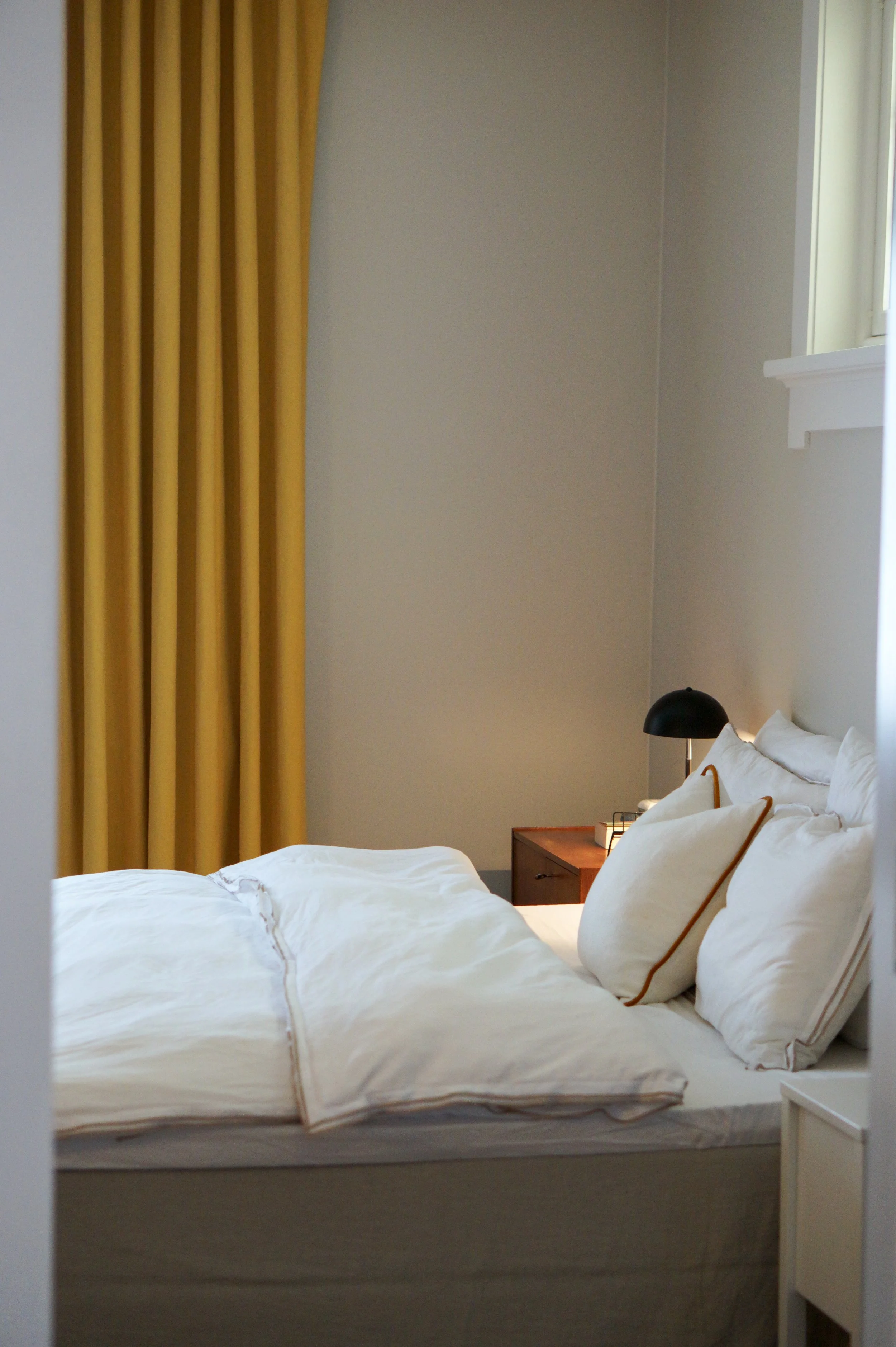 A neatly made bed with white bedding and pillows, next to a wooden nightstand with a black desk lamp, in a bedroom with beige walls and yellow curtains.