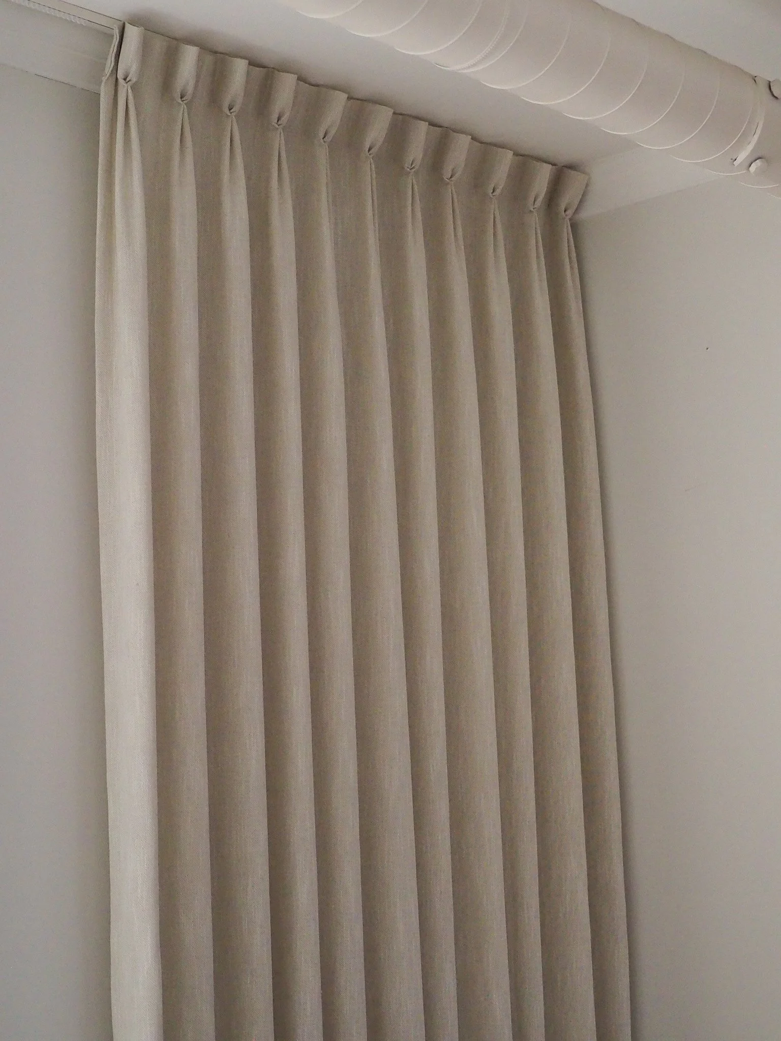 Beige curtains hanging from a curtain rod, mounted on a ceiling with white ductwork visible.