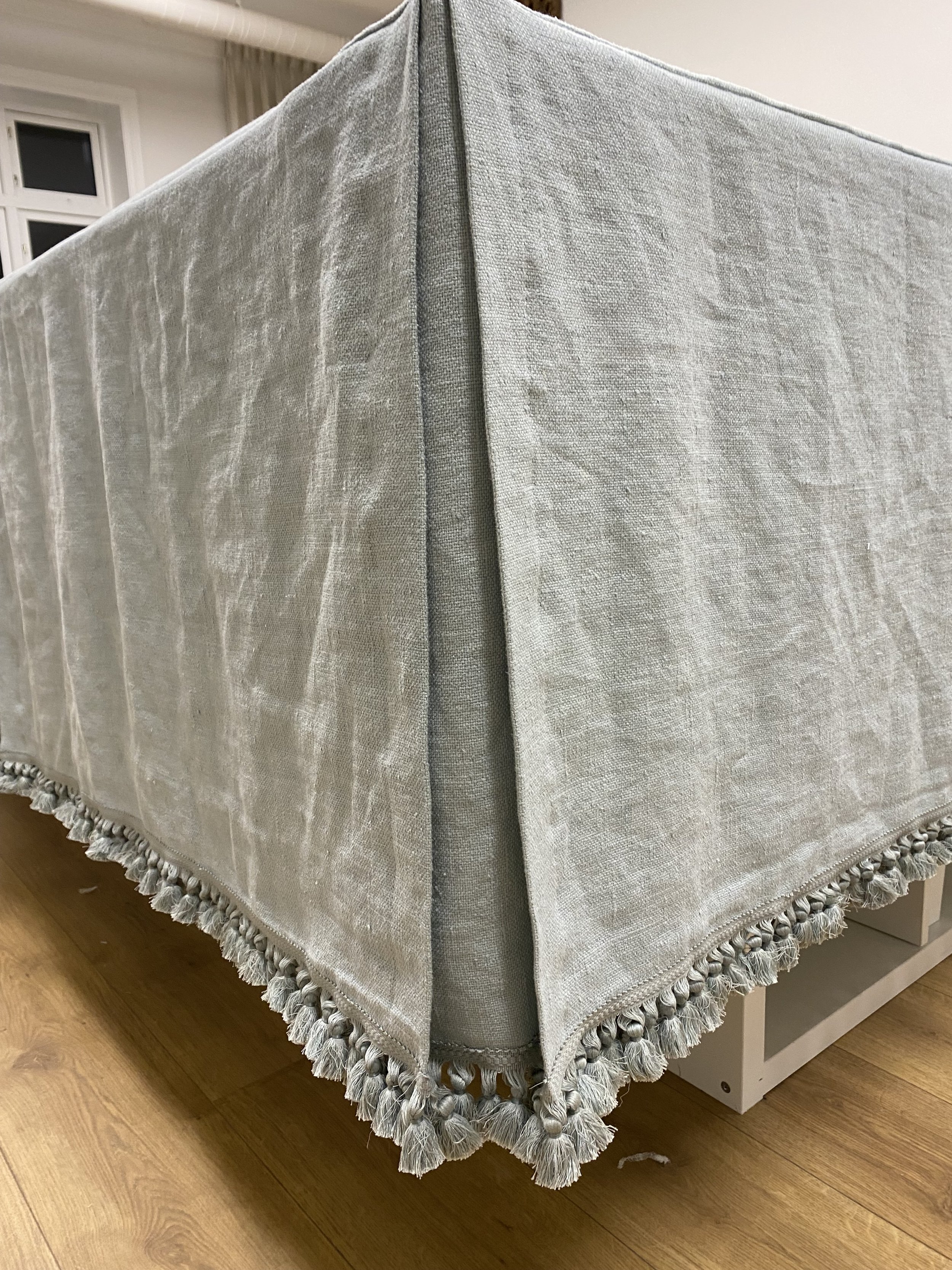 A beige or light gray tablecloth with textured pattern and decorative tassels hangs over a table, with a corner visible near a wooden floor.