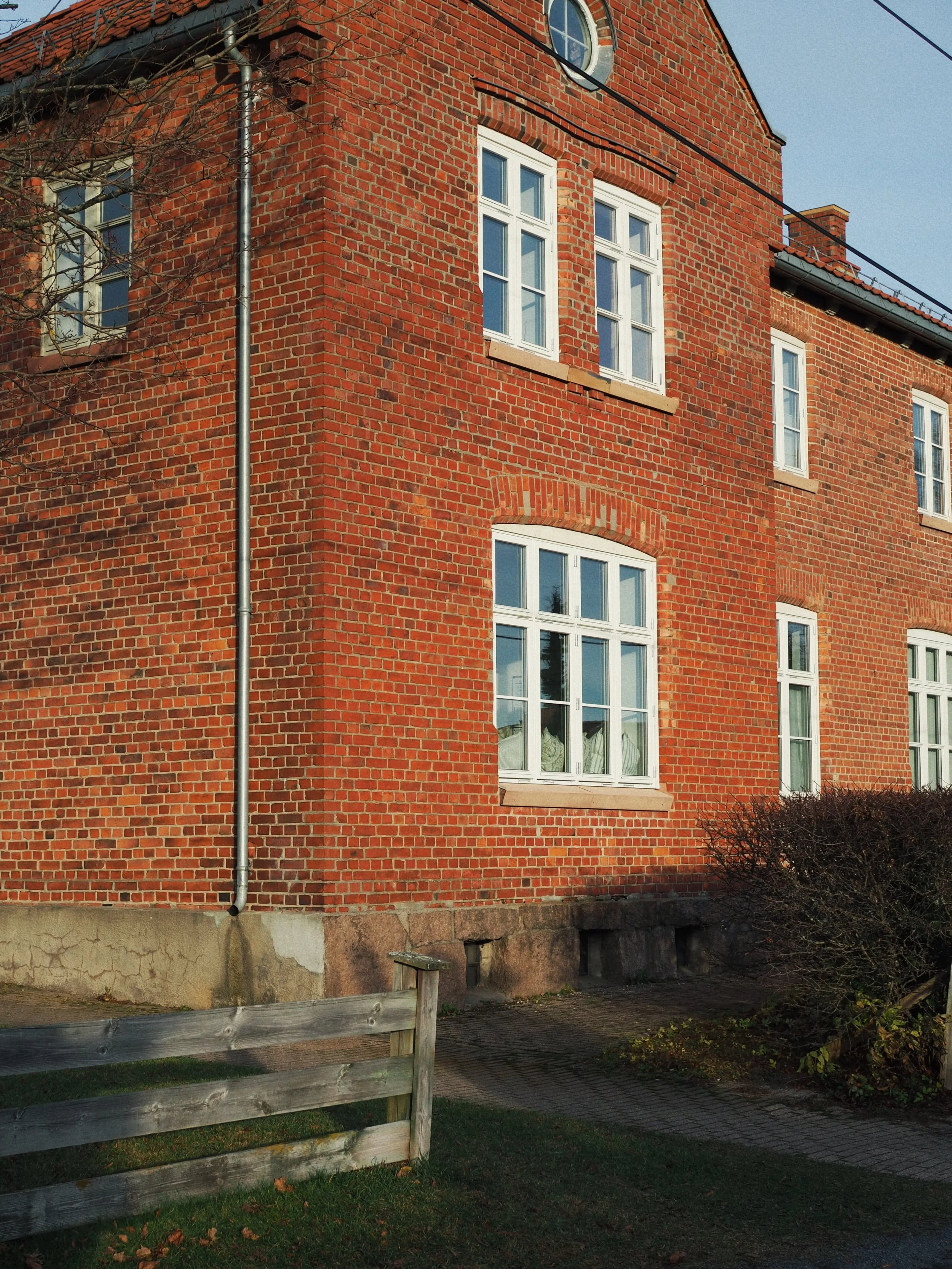 A red brick building with several white-framed windows, a small tree with no leaves, a grey gutter pipe running along the corner, and a wooden fence in the foreground. The sky is clear and blue.