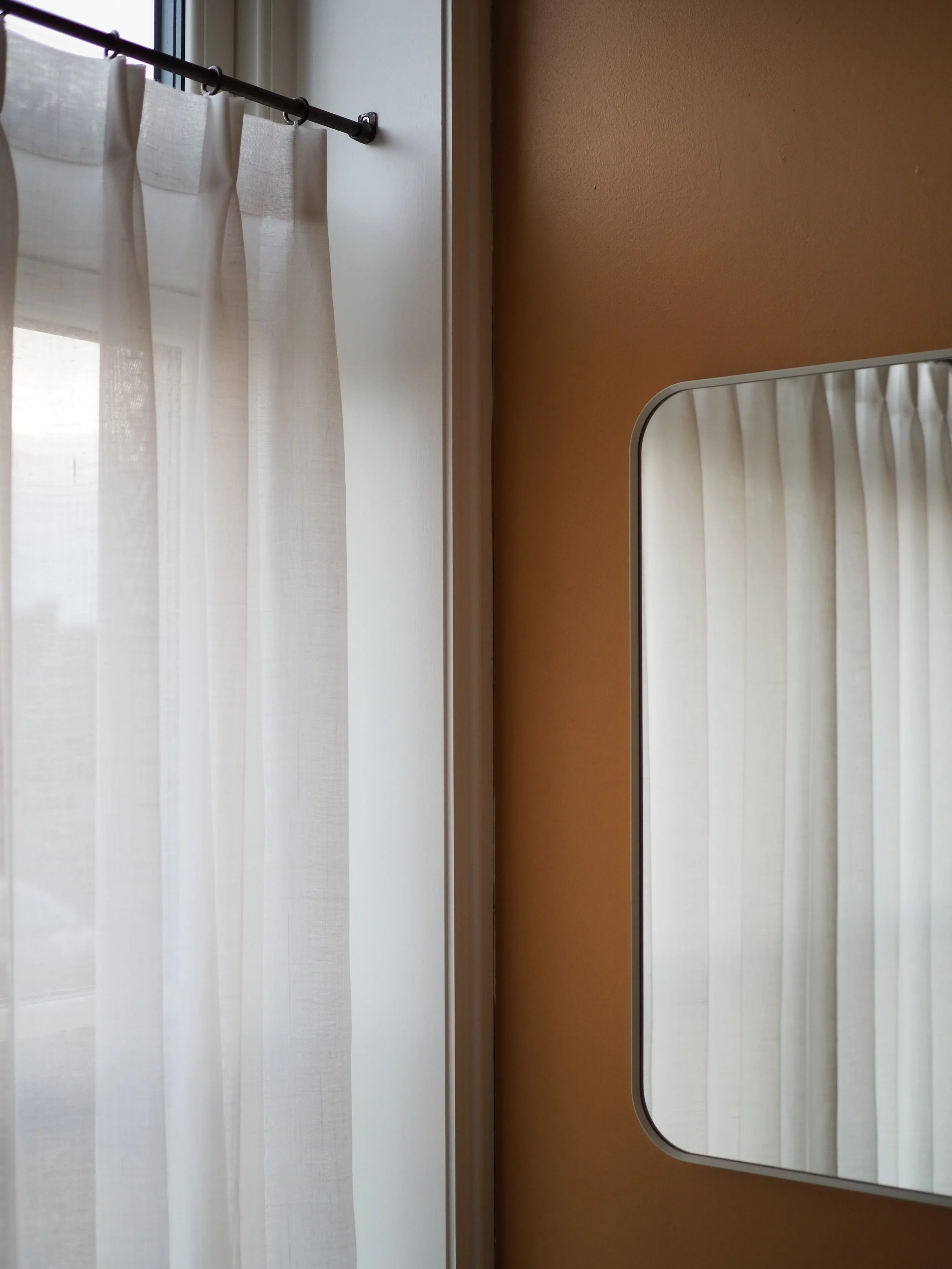 A room with beige walls, white curtains hanging from a black rod, and a rectangular mirror reflecting the curtains.