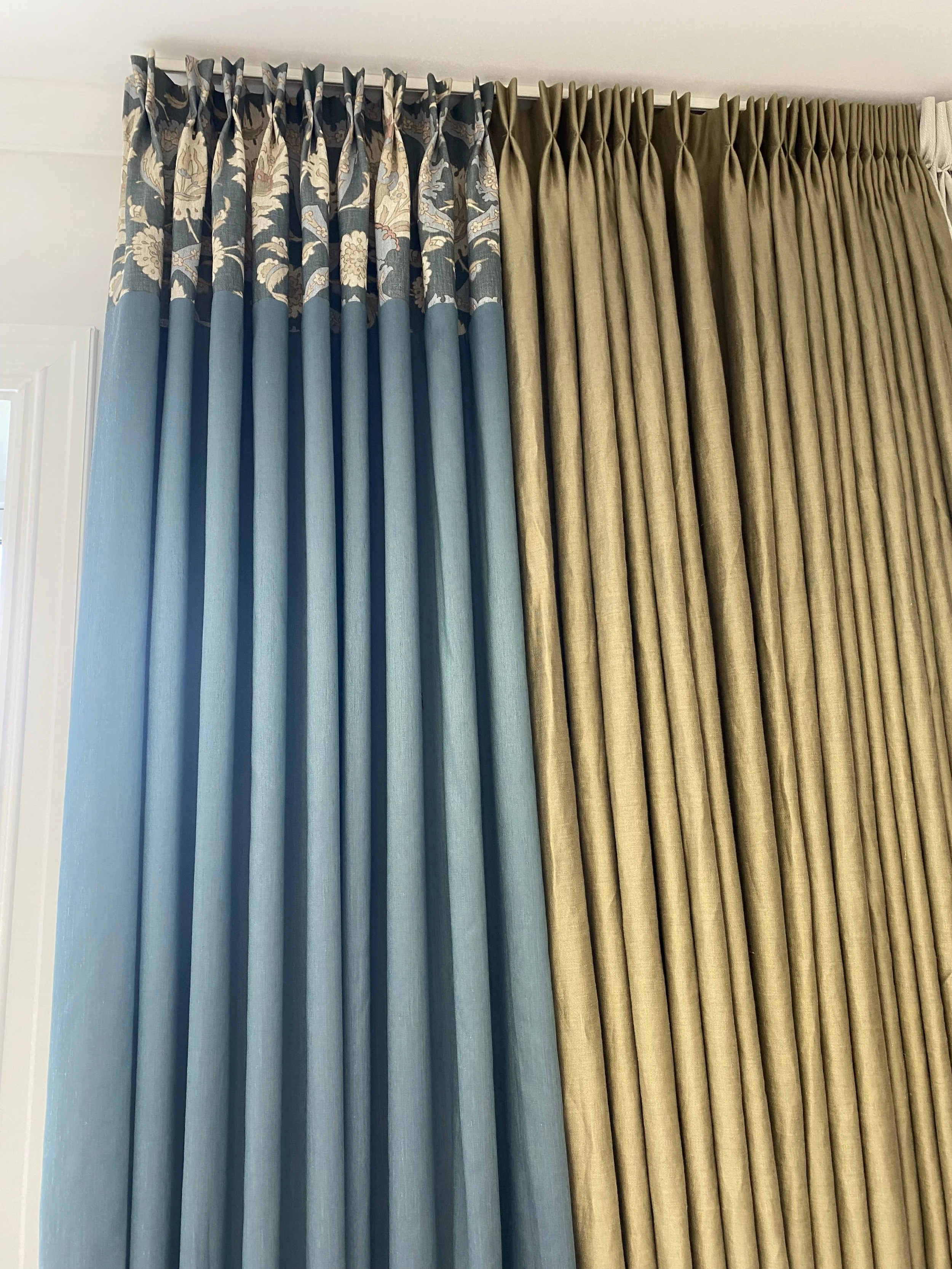 Two sets of curtains hanging side by side, one in light blue with a floral patterned top, and the other in goldish beige plain fabric.