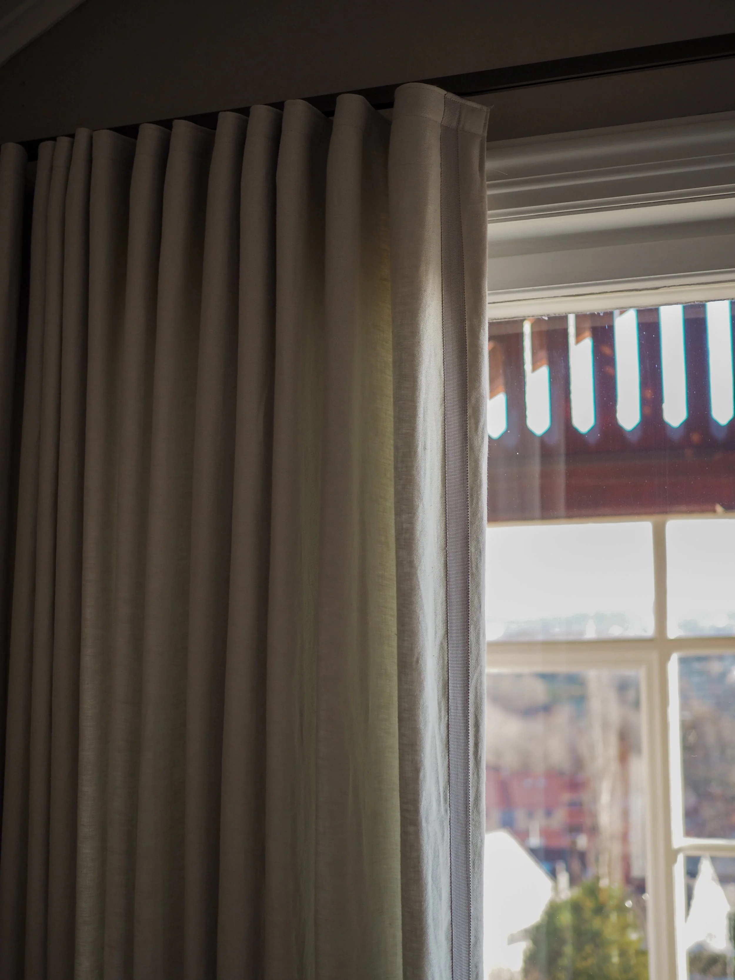 A close-up of beige curtains hanging by a window, with part of the window frame and outside view of buildings and a bright sky visible.