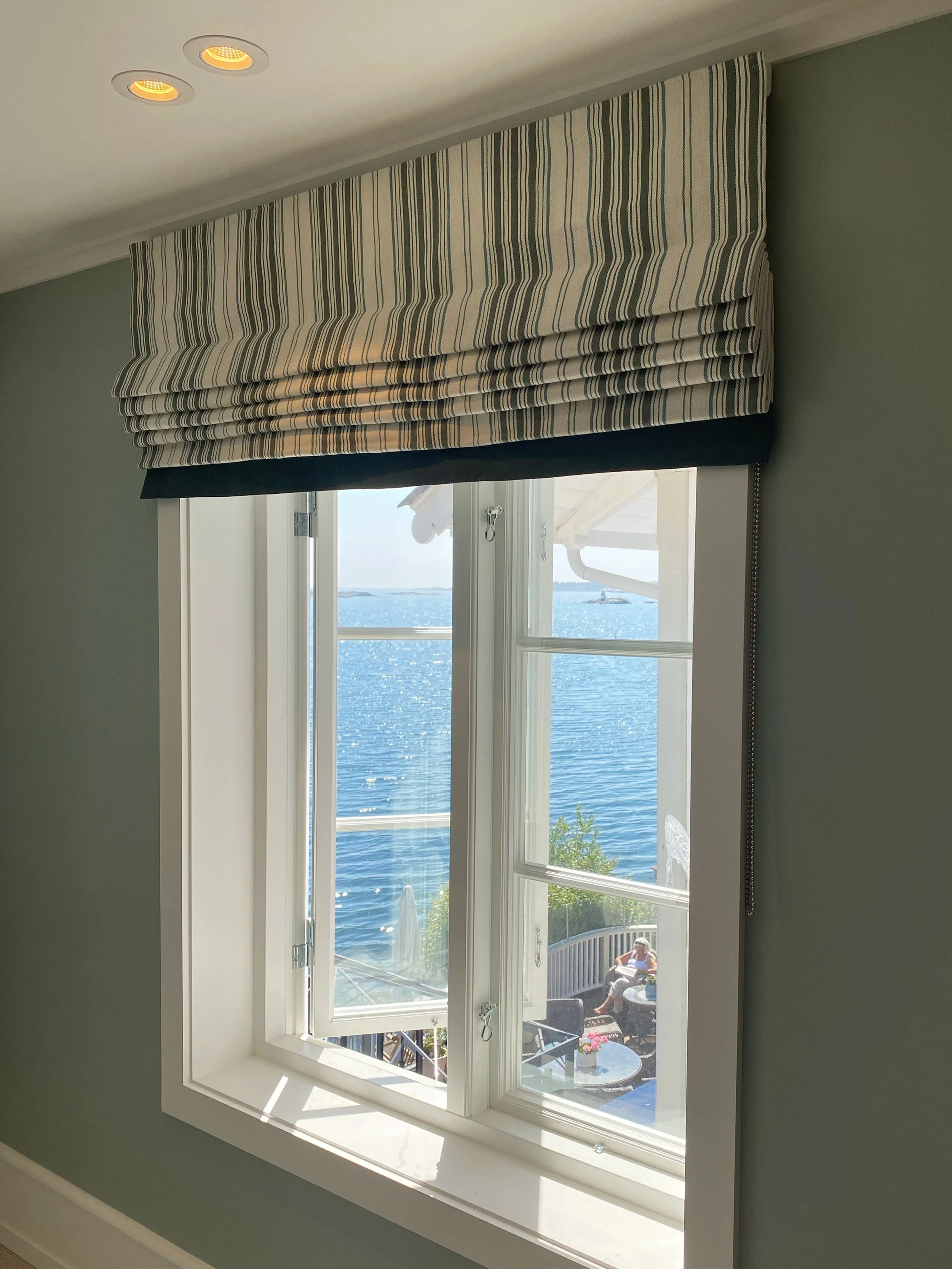 A window with striped valances overlooking a patio by the water, with a woman sitting outside.